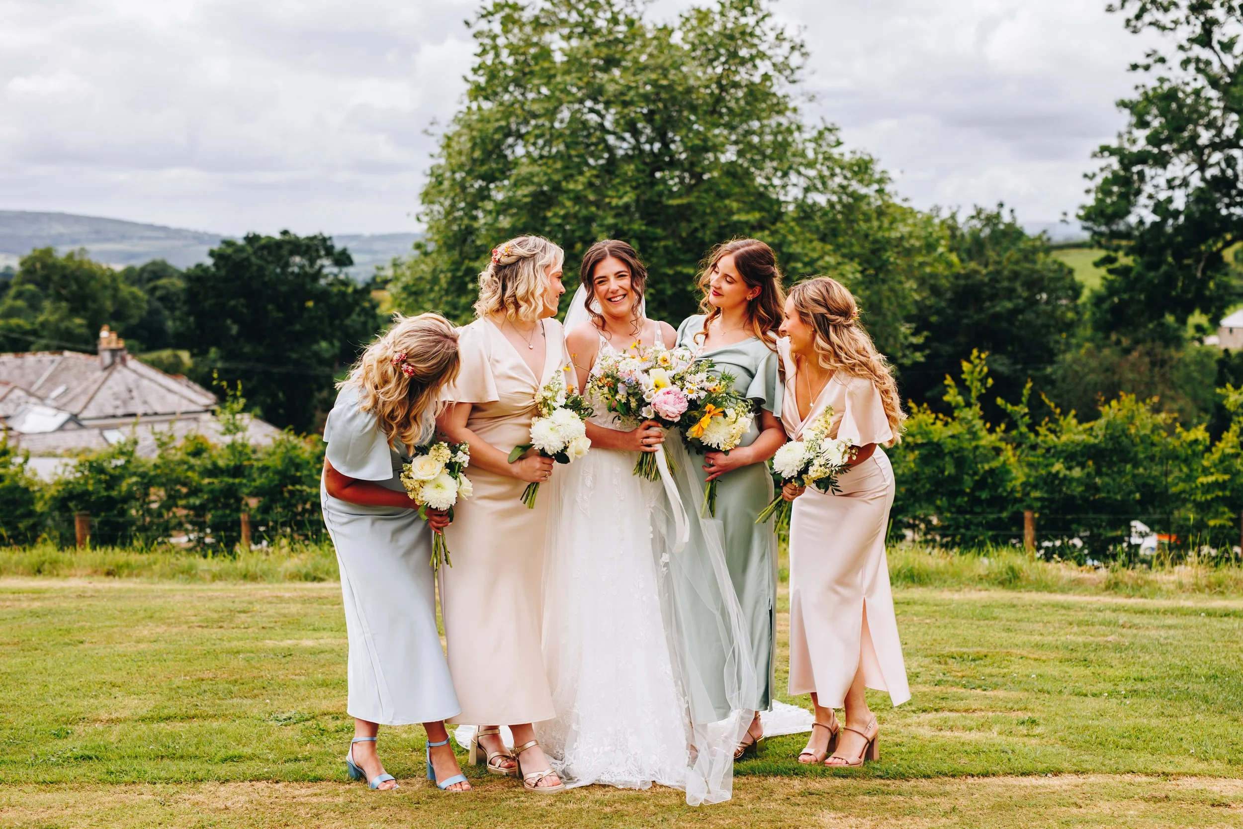 Bride with four bridesmaids holding bouquets, standing on grass outdoors with trees and hills in the background.