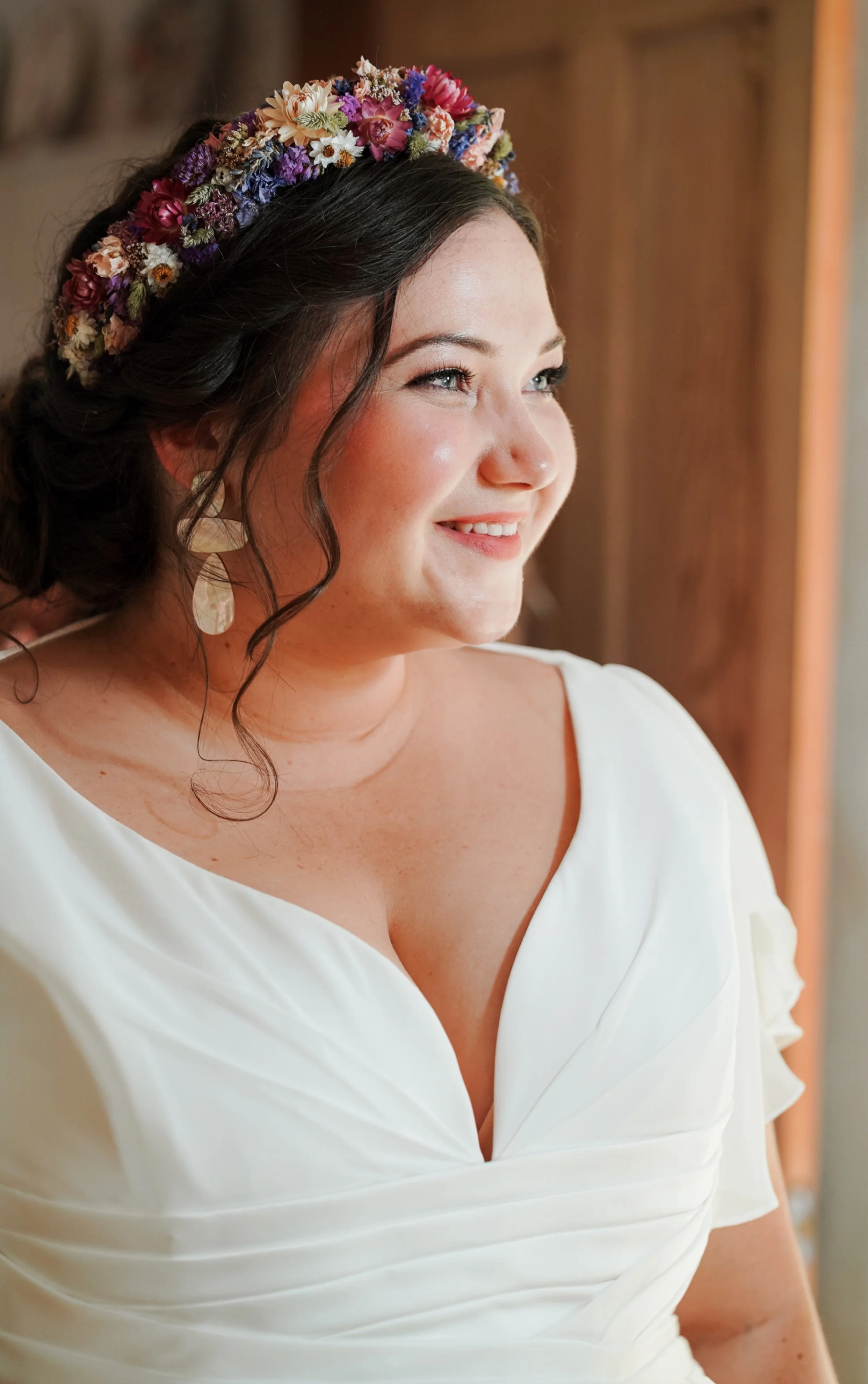 A woman with dark hair wearing a flower crown, white dress, and earrings, smiling and looking to her right.