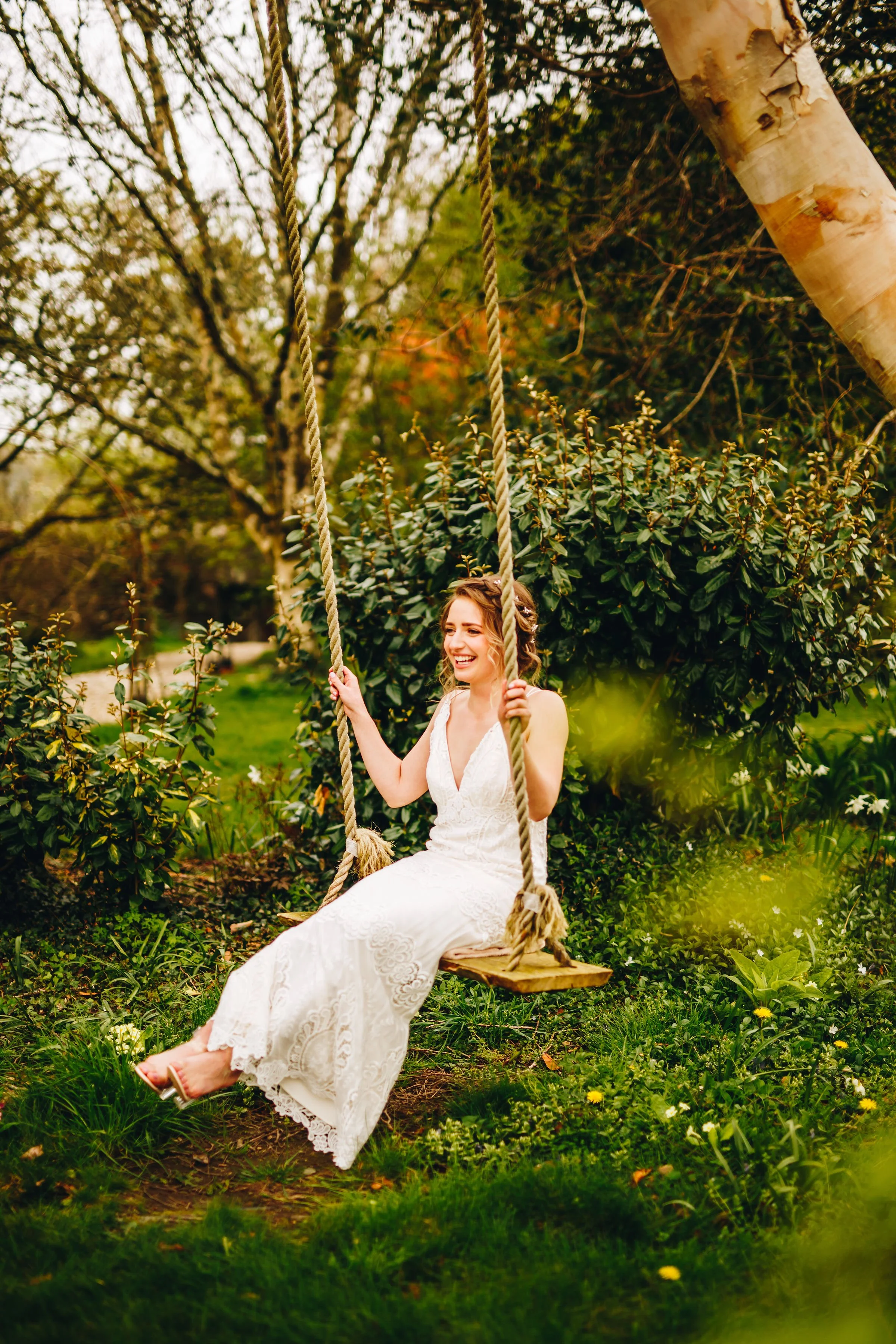 A woman in a white dress swinging on a wooden swing in a lush garden with green grass and trees.