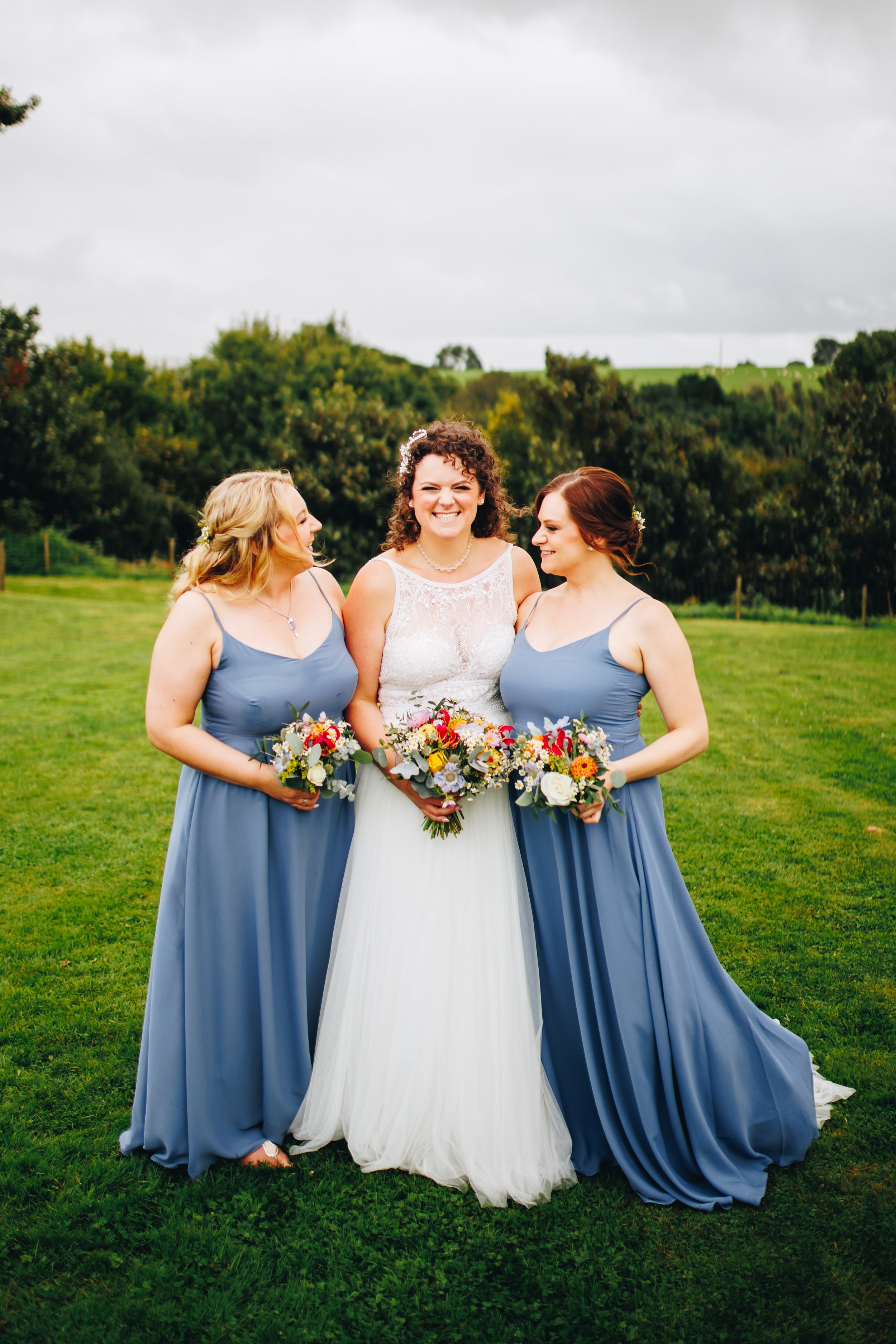 A bride with curly hair and two bridesmaids in blue dresses holding bouquets on a grass field with trees and cloudy sky in the background.