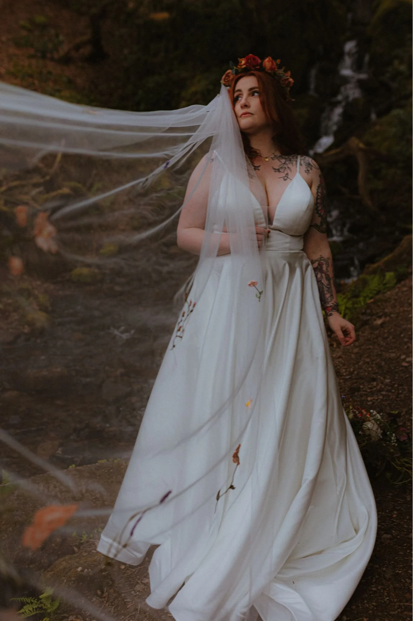 A woman wearing a white wedding dress with tattoos, a flower crown, and veil stands in a natural outdoor setting near a small waterfall.