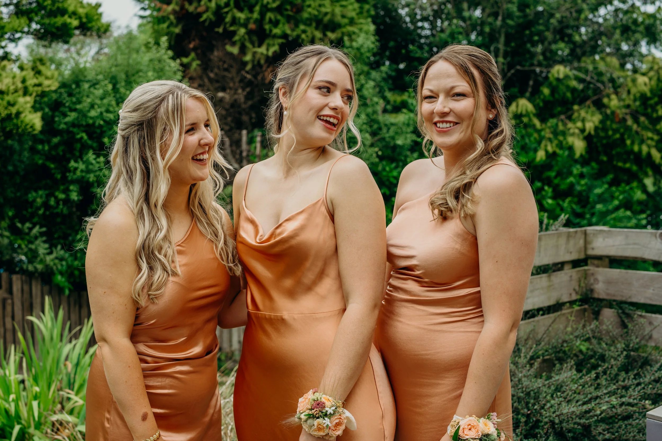 Three women in matching peach satin dresses standing outside, smiling and talking, with greenery in the background.