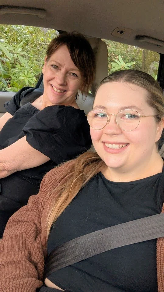 Two women smiling inside a car with greenery visible through the window.