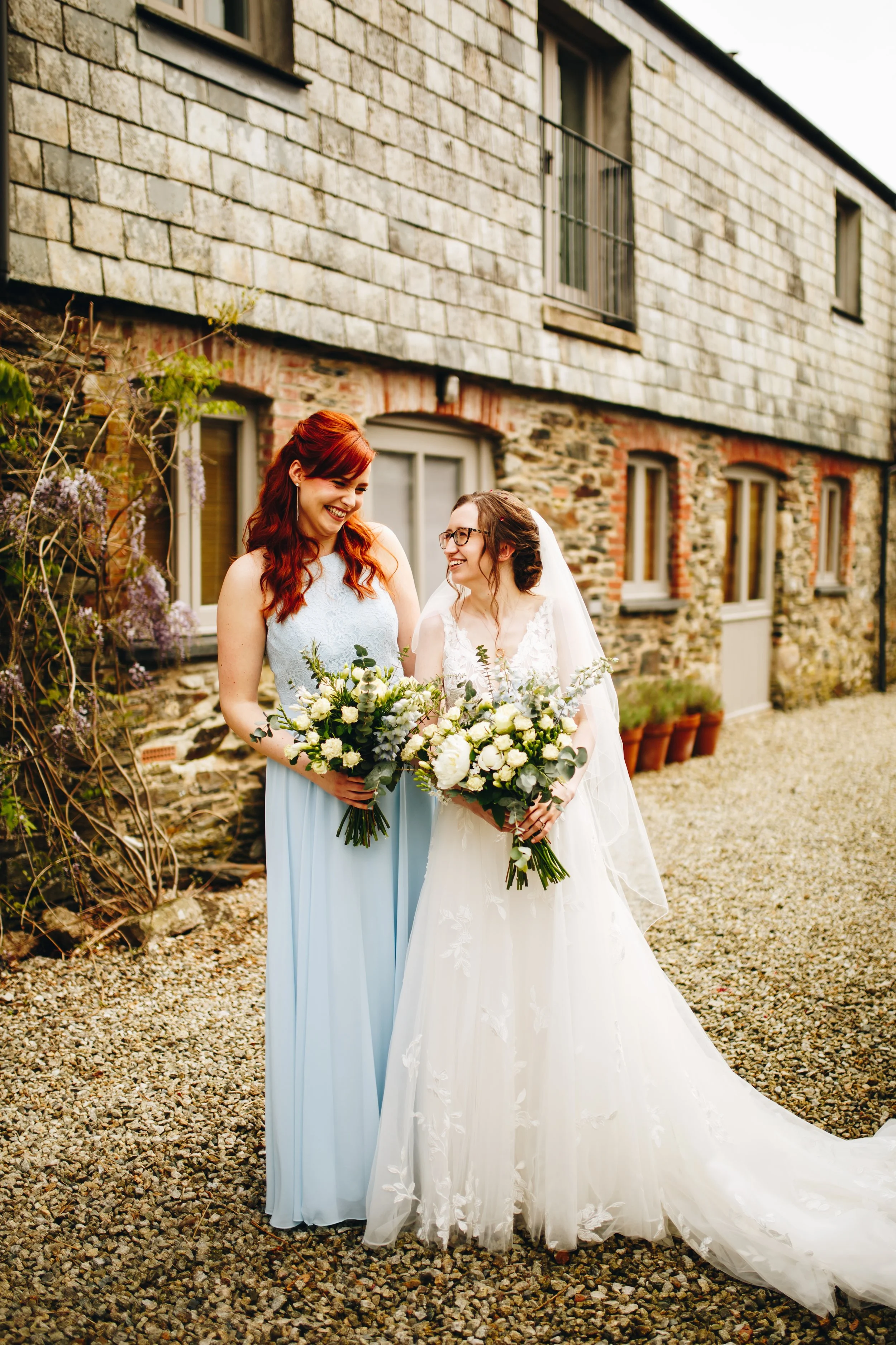 Two women, one in a wedding dress and one in a light blue bridesmaid dress, holding bouquets of white flowers, smiling and looking at each other outdoors in front of a stone and brick building.