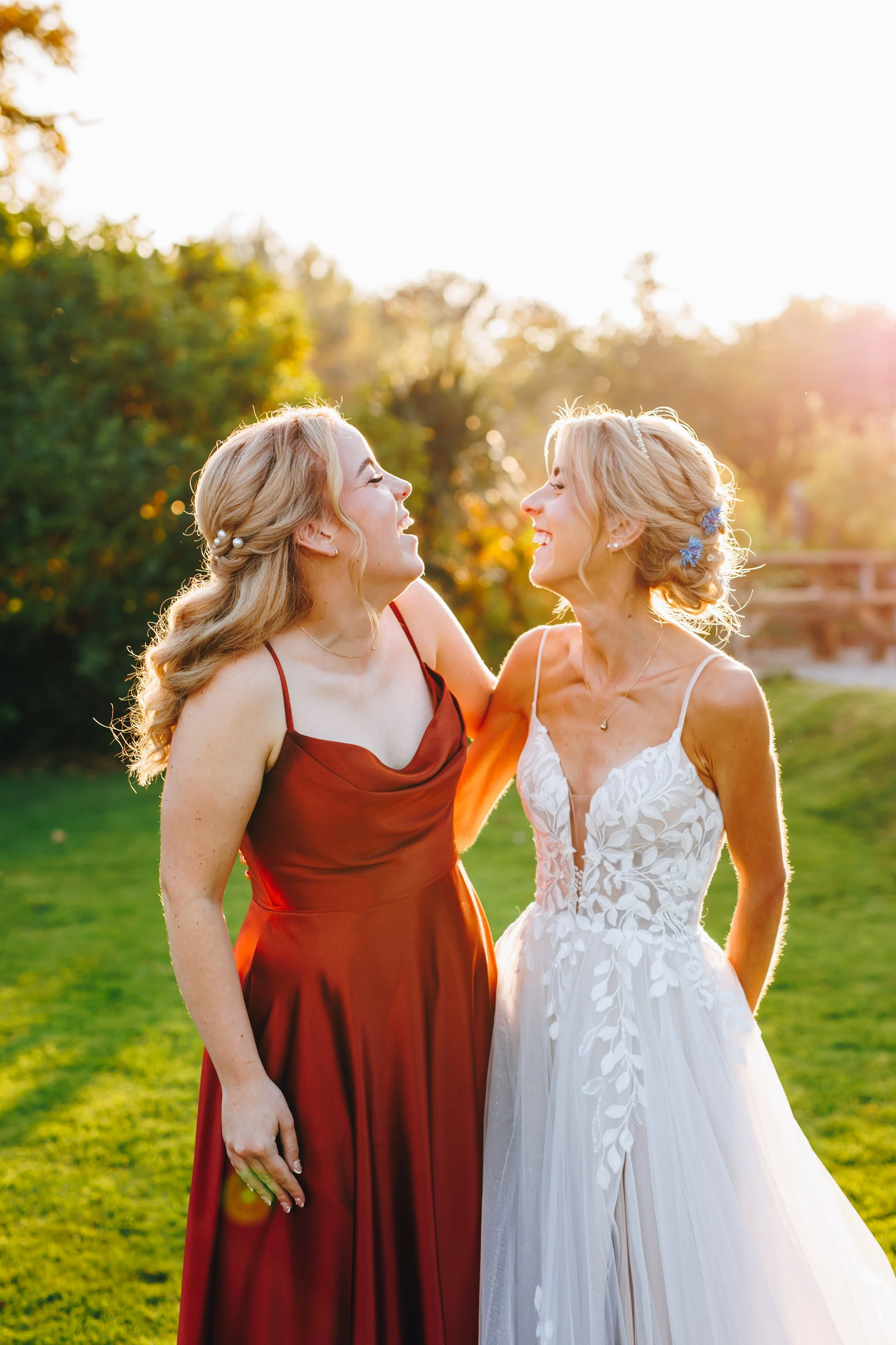 Two women, one in a dark red dress and the other in a white wedding gown, smiling and looking at each other outdoors in a grassy area during sunset.