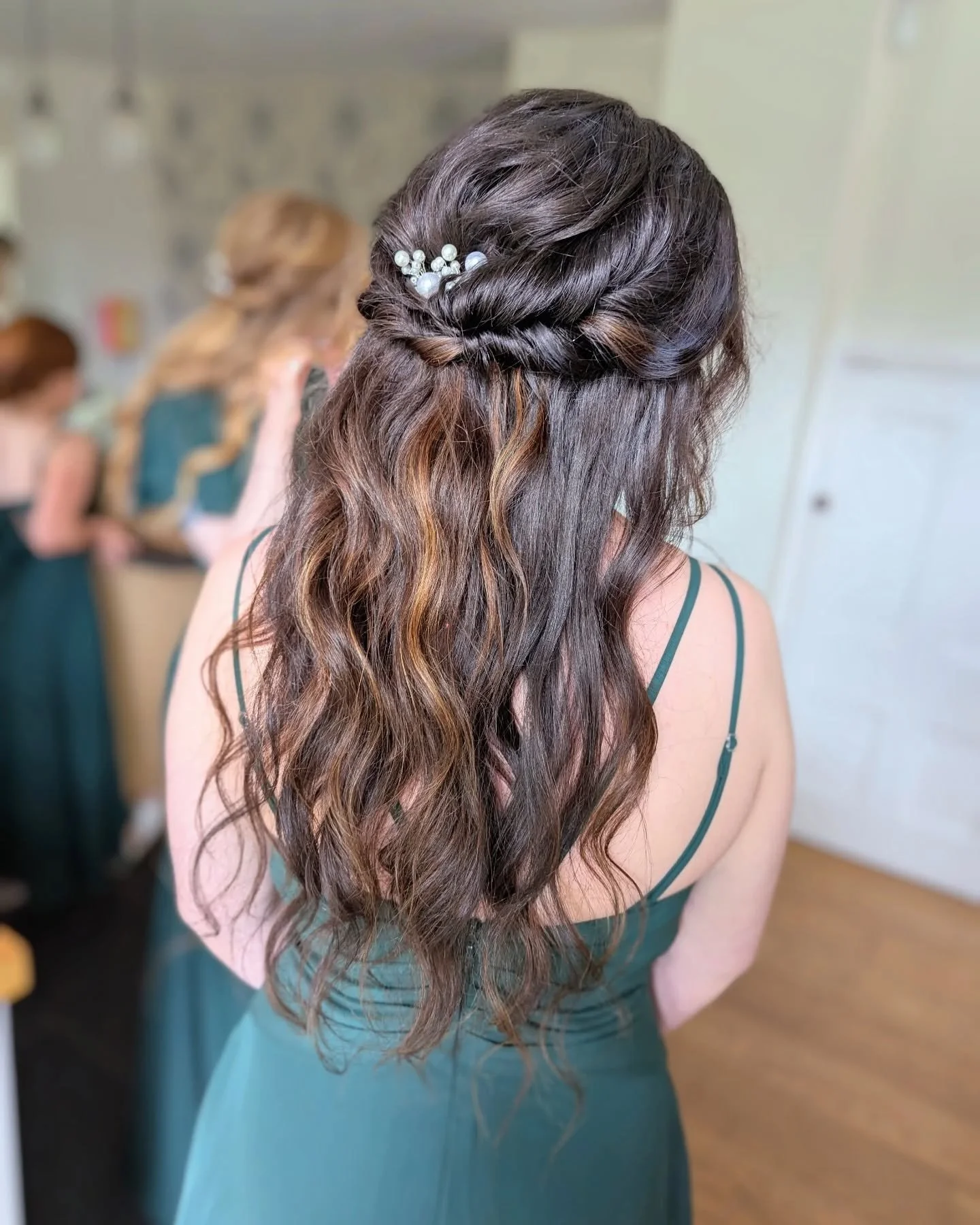 Back view of a woman with long wavy brown hair styled with a twist braid and pearl hair clip, wearing a teal dress with thin straps, in a room with other women in similar dresses.