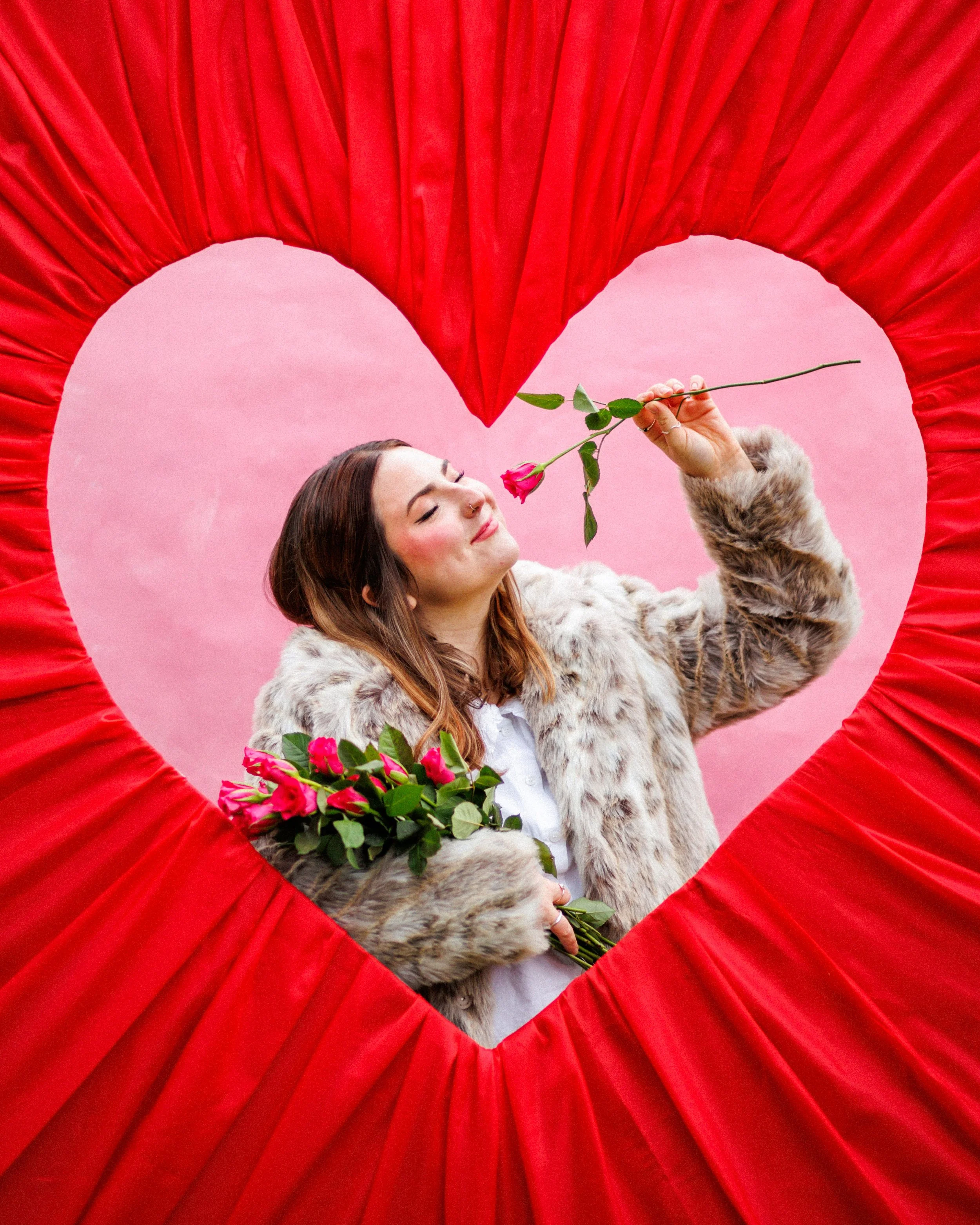 A woman holding pink roses and smiling, seen through a heart-shaped opening in a red curtain, with a pink background.