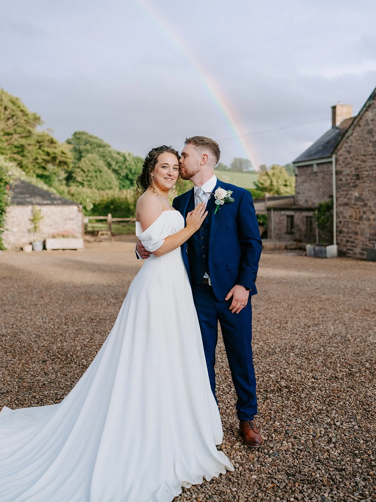 A bride and groom stand outdoors on a gravel area with green trees and a stone house in the background, under a rainbow in a cloudy sky. The bride is wearing a white off-the-shoulder wedding dress, and the groom is in a dark blue suit with a white sh