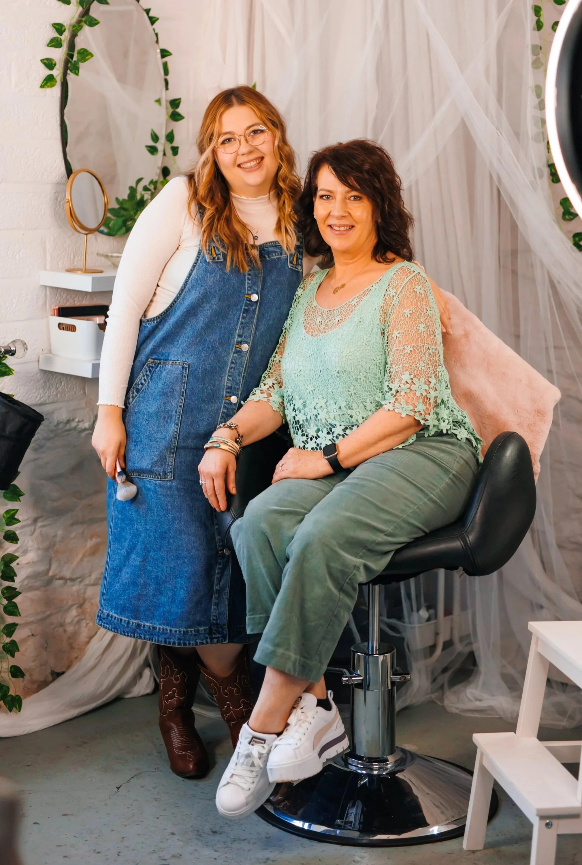 Two women smiling and posing indoors, with one seated on a stool and the other standing beside her. The seated woman has shoulder-length dark hair and is wearing a light green lace top and green pants, while the standing woman has long hair, glasses, and is wearing a white long-sleeve top under a denim overall dress.