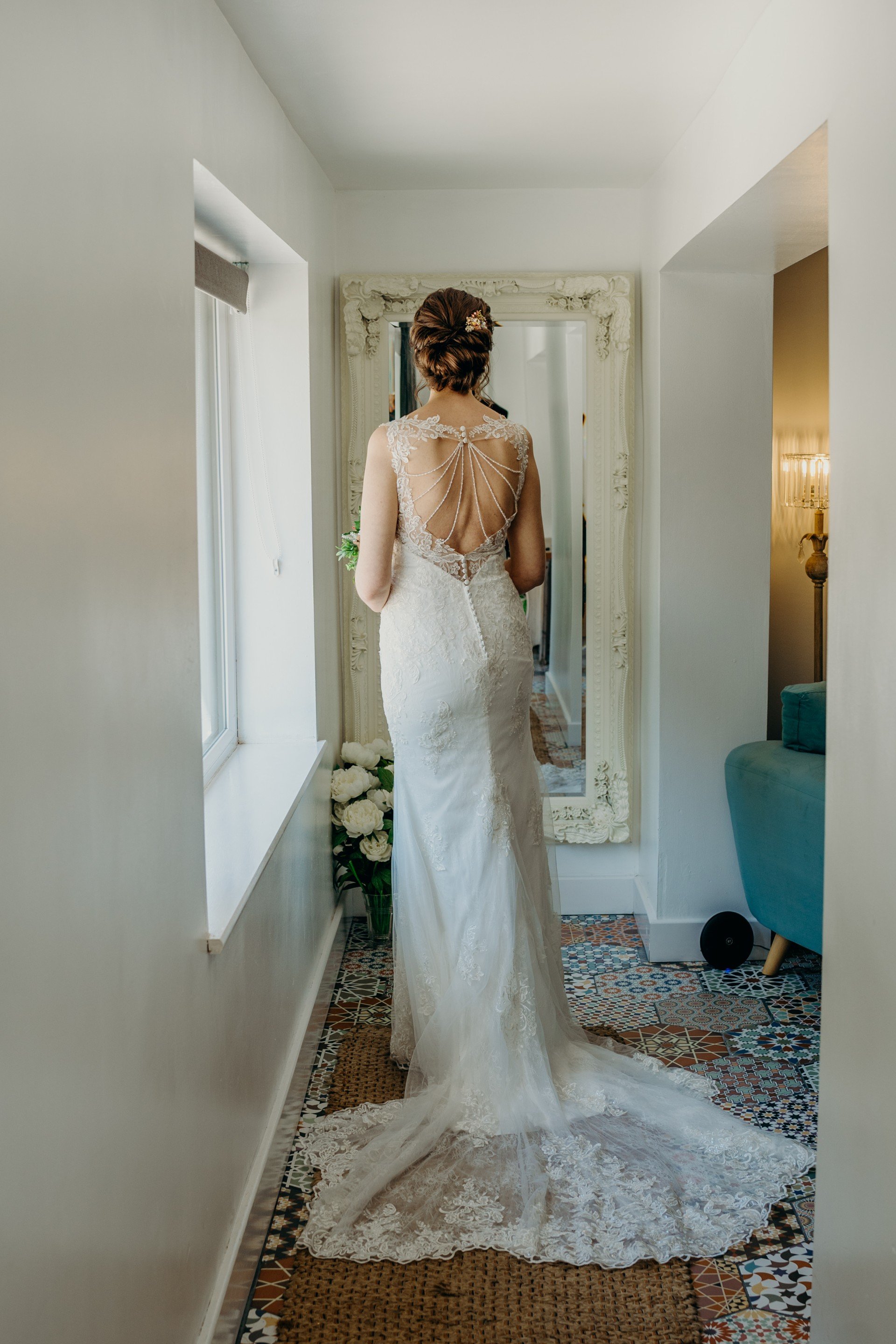 Bride in a wedding dress with a lace back detail, standing in front of a mirror in a brightly lit room, with floral decor nearby.
