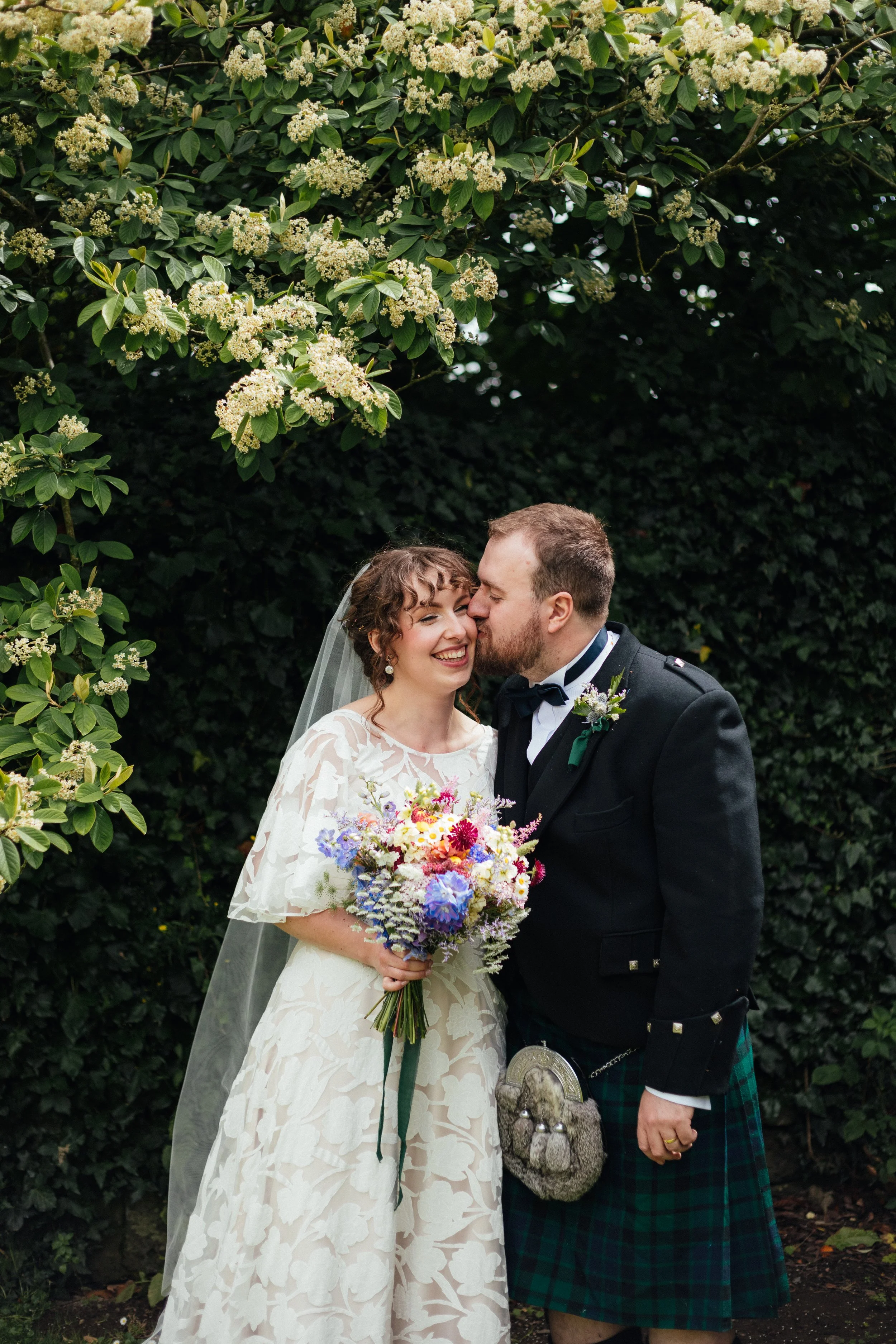 A bride and groom on their wedding day, standing under greenery with white flowers. The bride is holding a colorful bouquet and wearing a lace wedding dress with a veil. The groom is dressed in a traditional Scottish kilt and jacket, kissing the bride.
