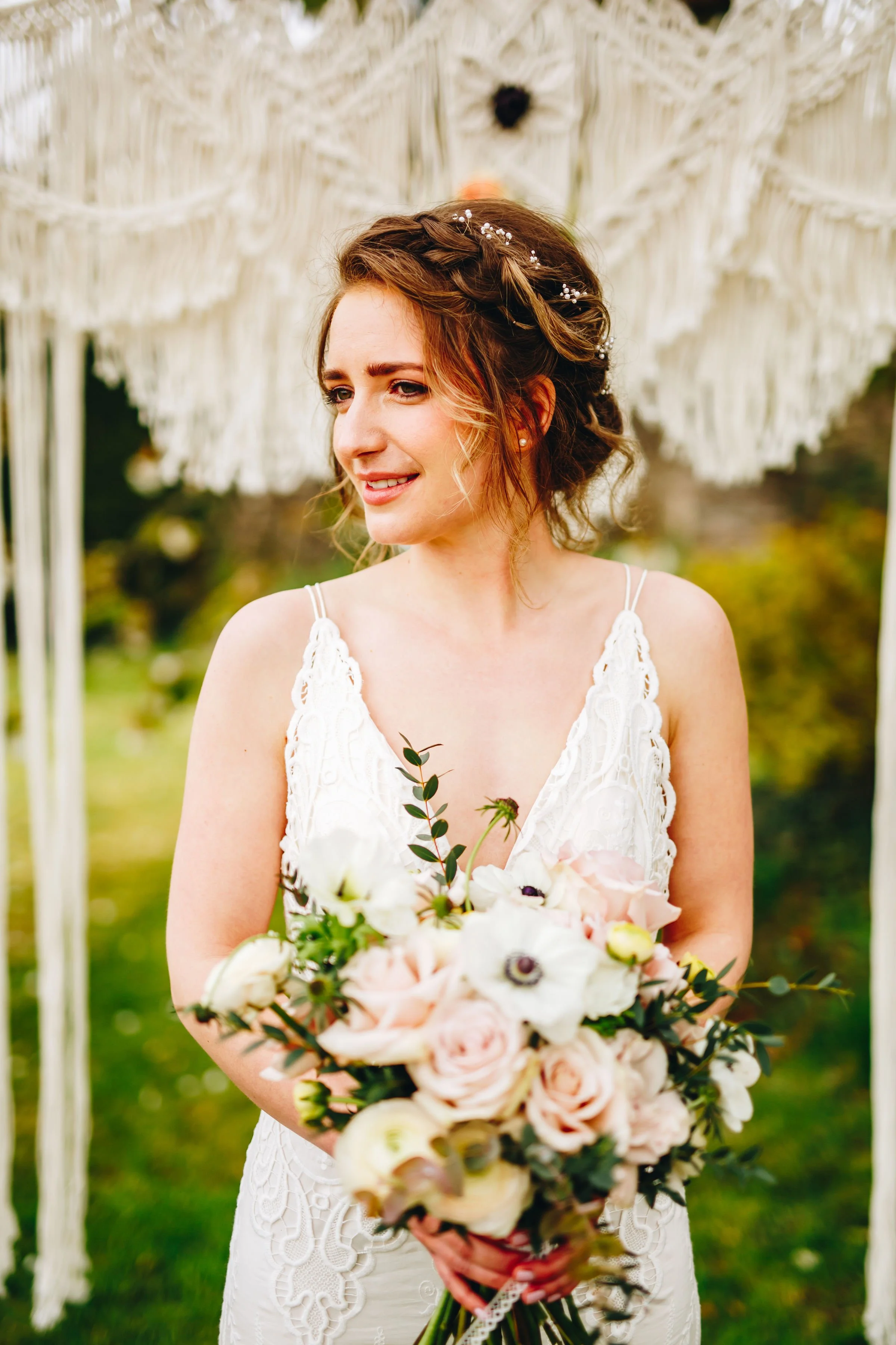 A woman with braided hair and a white lace dress holding a bouquet of pink and white flowers, standing outdoors under a macrame decoration.