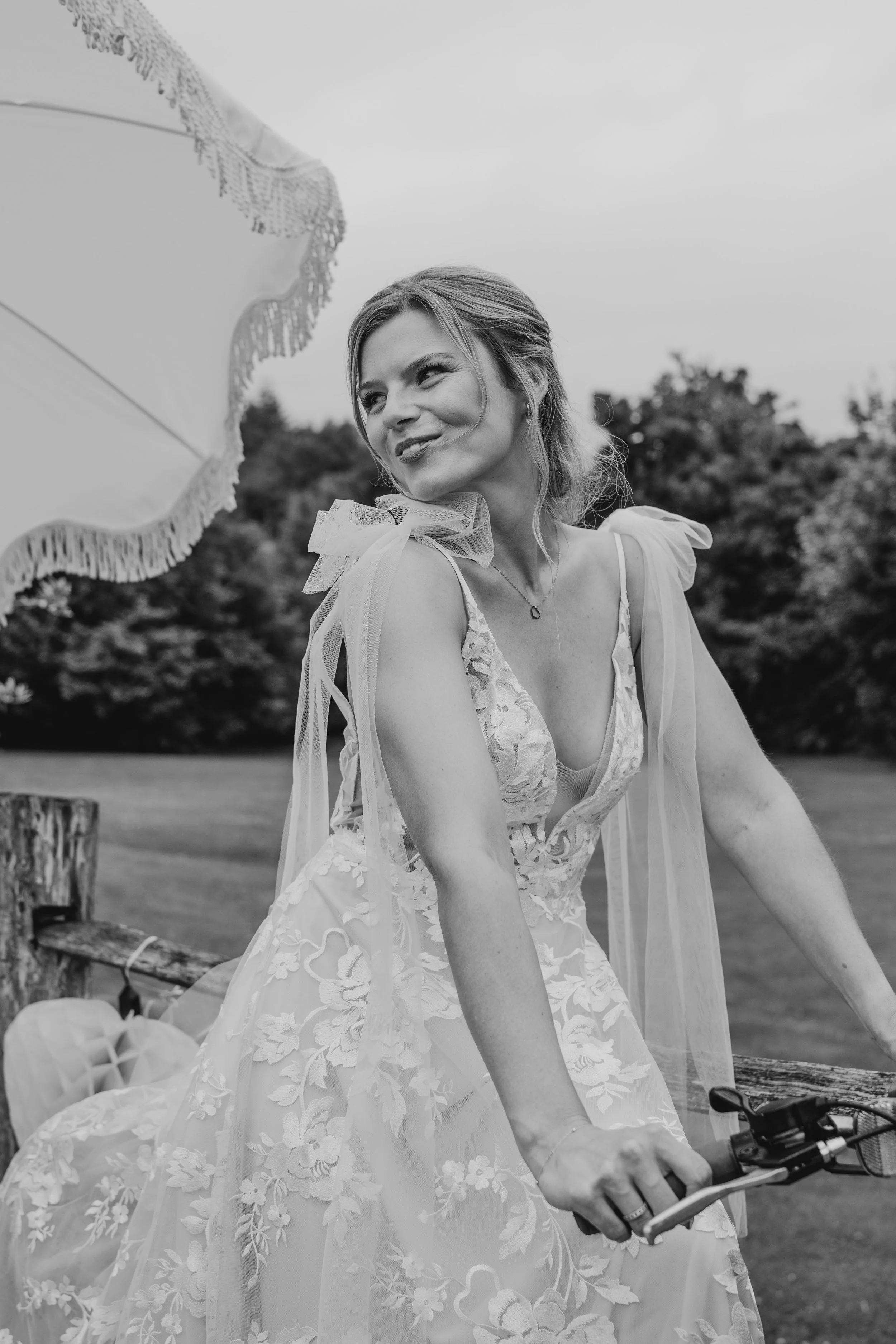 A woman in a wedding dress riding a bicycle outdoors, smiling and looking to the side, with trees in the background.