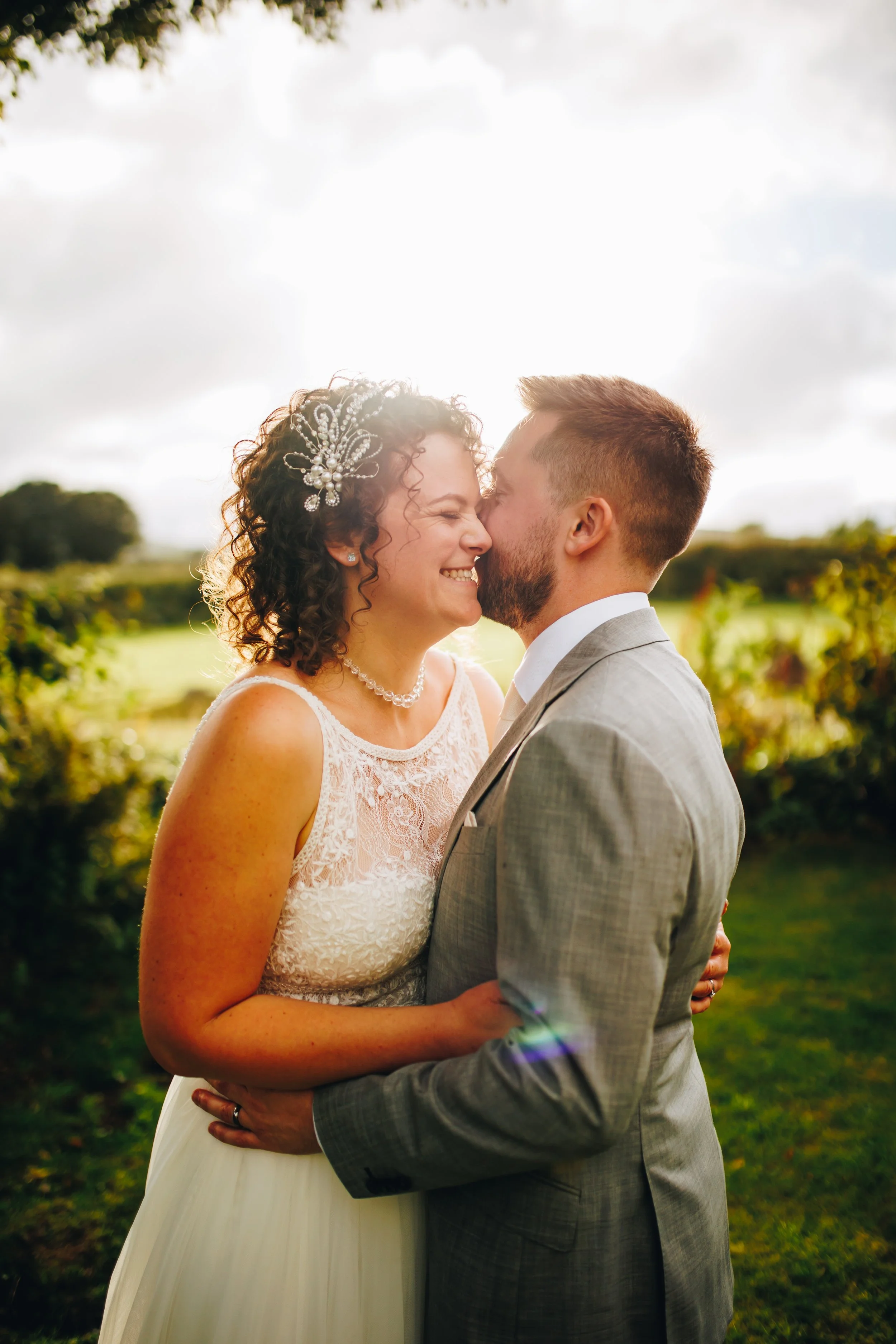 A bride and groom smiling and sharing a kiss outdoors on their wedding day, with a vineyard in the background and sunlight shining behind them.