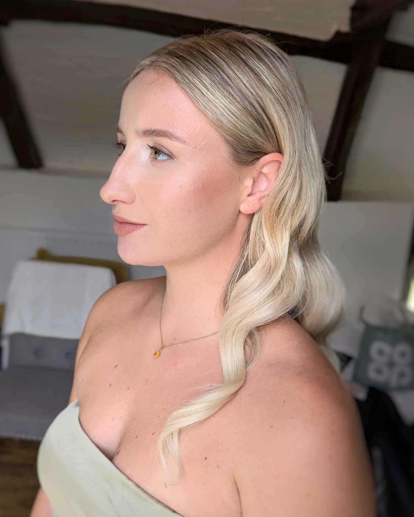 Side profile of a woman with blonde wavy hair and light skin, wearing a strapless top and a delicate necklace, in a room with dark wooden beams on the ceiling.