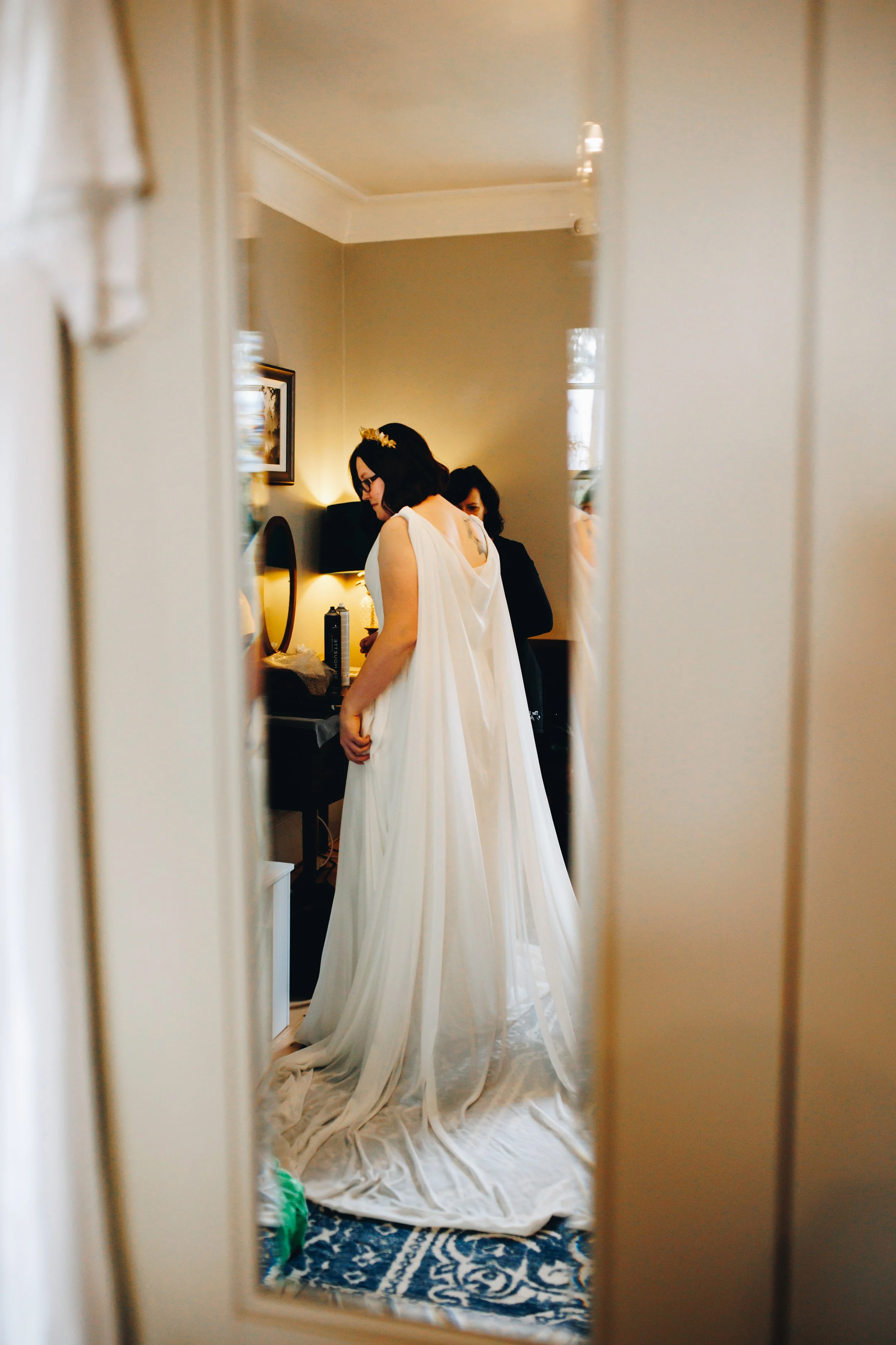 A woman in a white wedding dress with a floral headpiece is trying on her wedding gown, seen through a mirror, while a person behind her assists with the dress.
