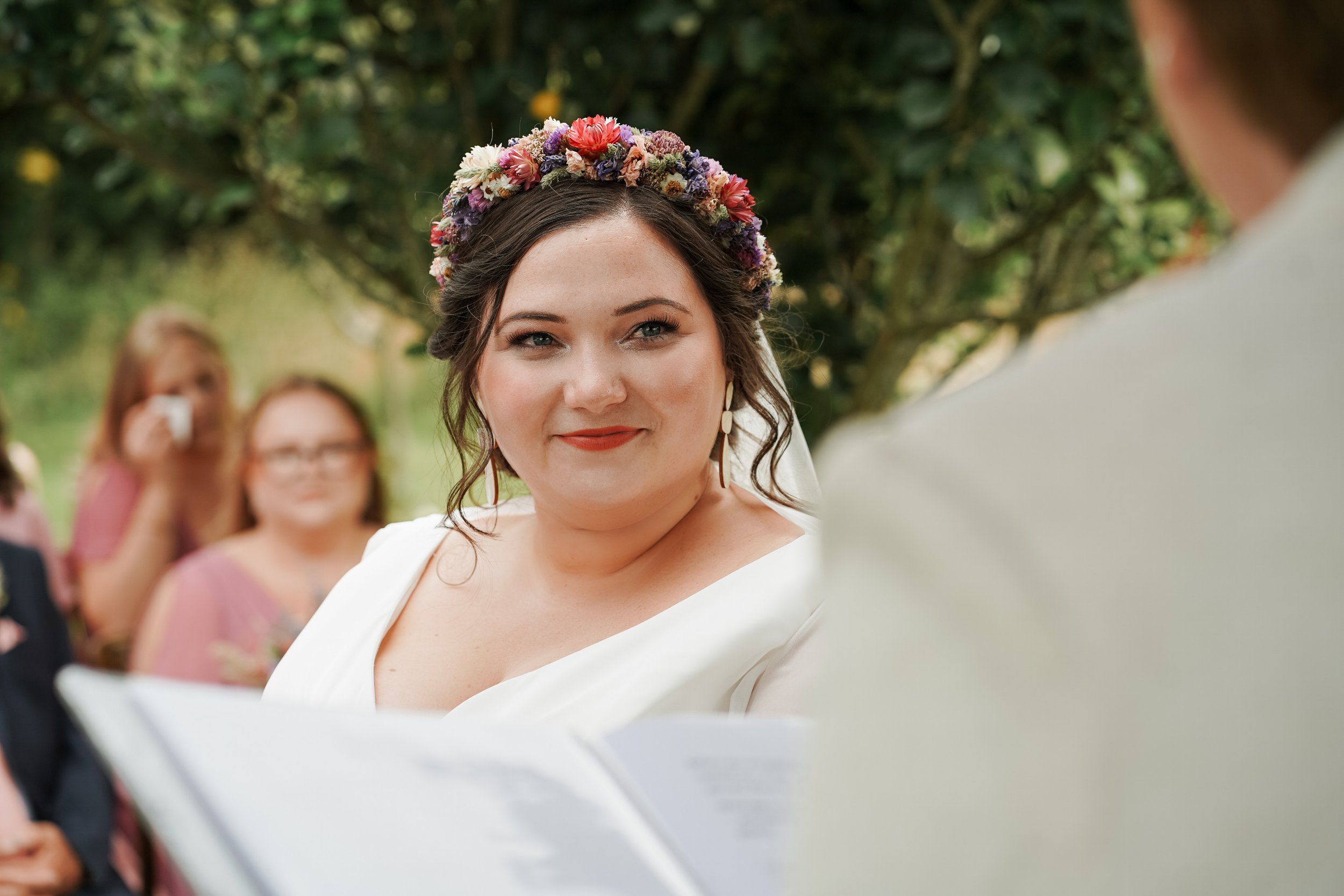 Bride with dark hair wearing a floral crown and white dress, smiling during her wedding ceremony outdoors, blurred guests in background.