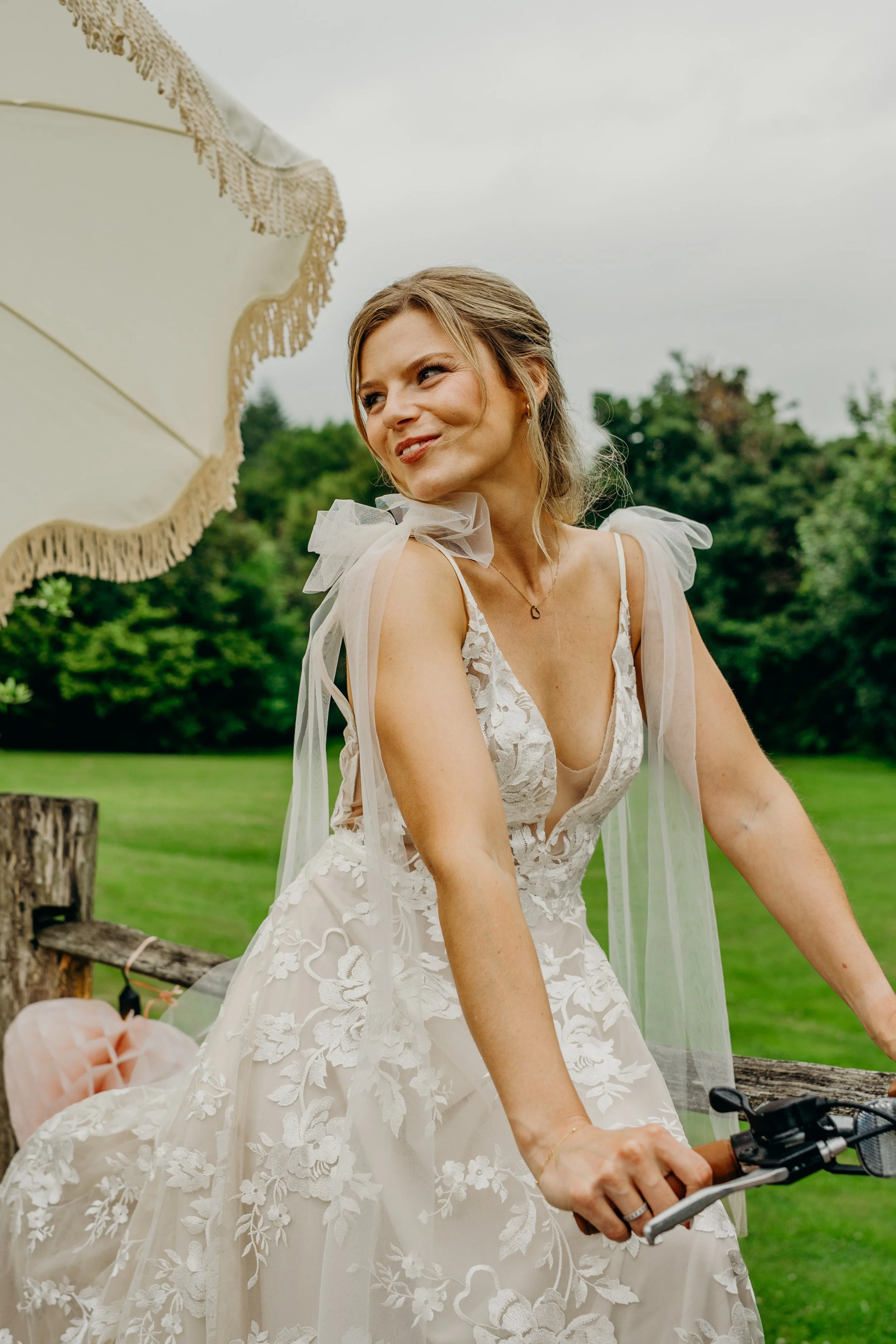 A woman in a white lace wedding dress with tulle shoulder accents, riding a bicycle outdoors on a cloudy day, with trees and green grass in the background.
