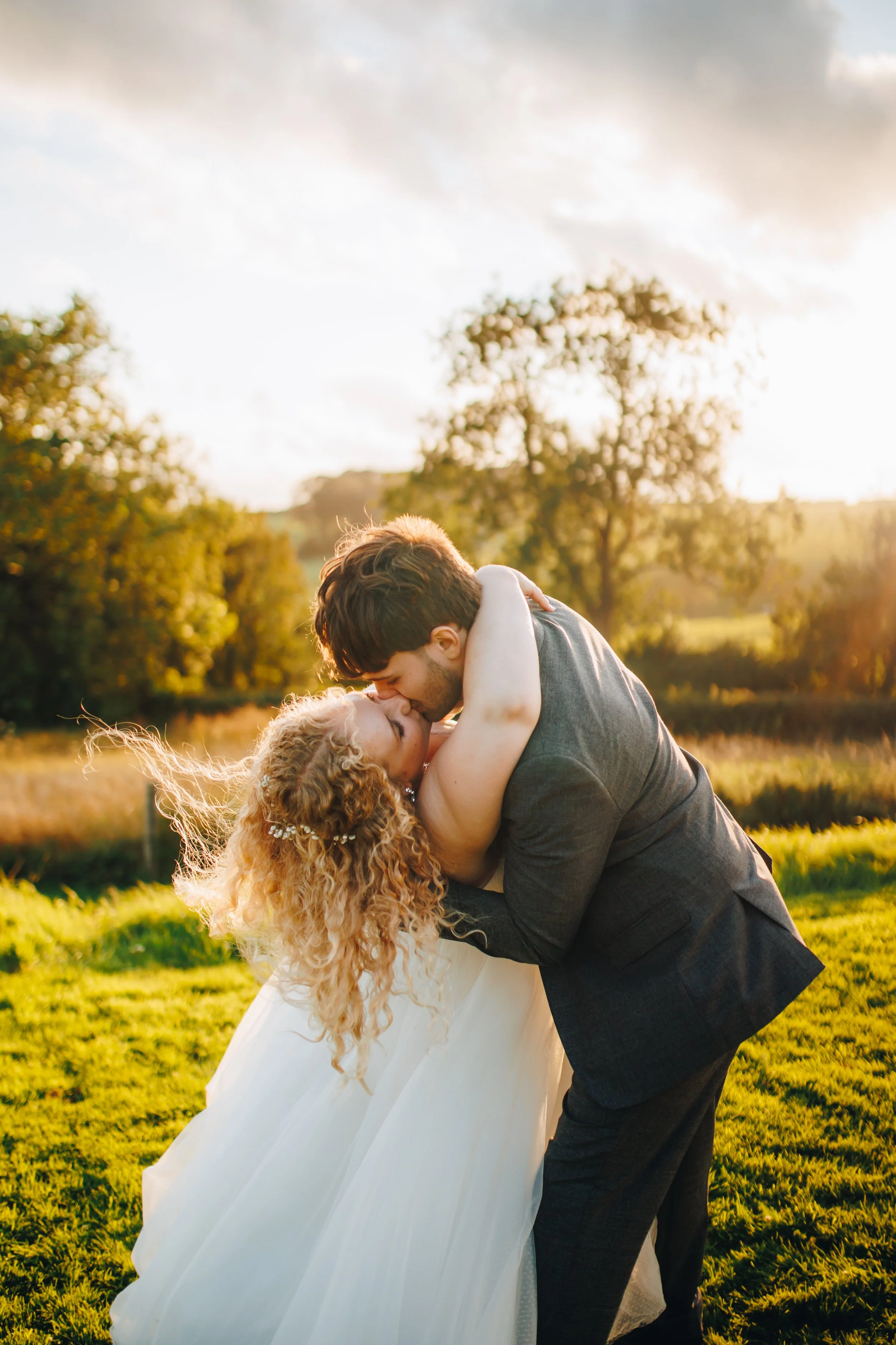 A couple sharing a romantic kiss outdoors during sunset, with the woman in a white wedding dress and the man in a dark suit, surrounded by a green field and trees.