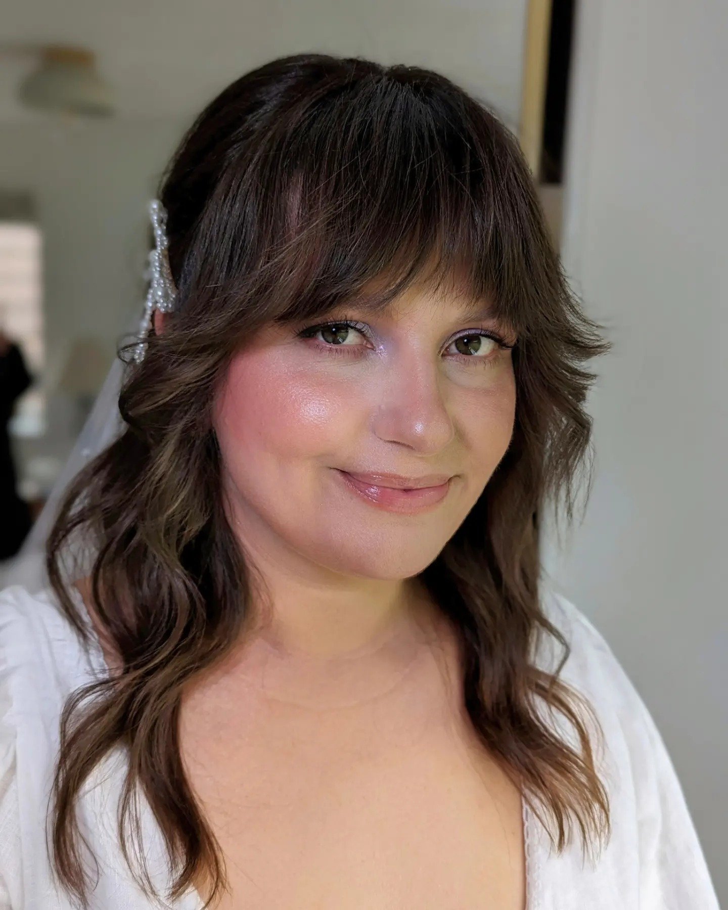 A woman with wavy brown hair, light makeup, and a white top, smiling gently at the camera, with a blurred indoor background.