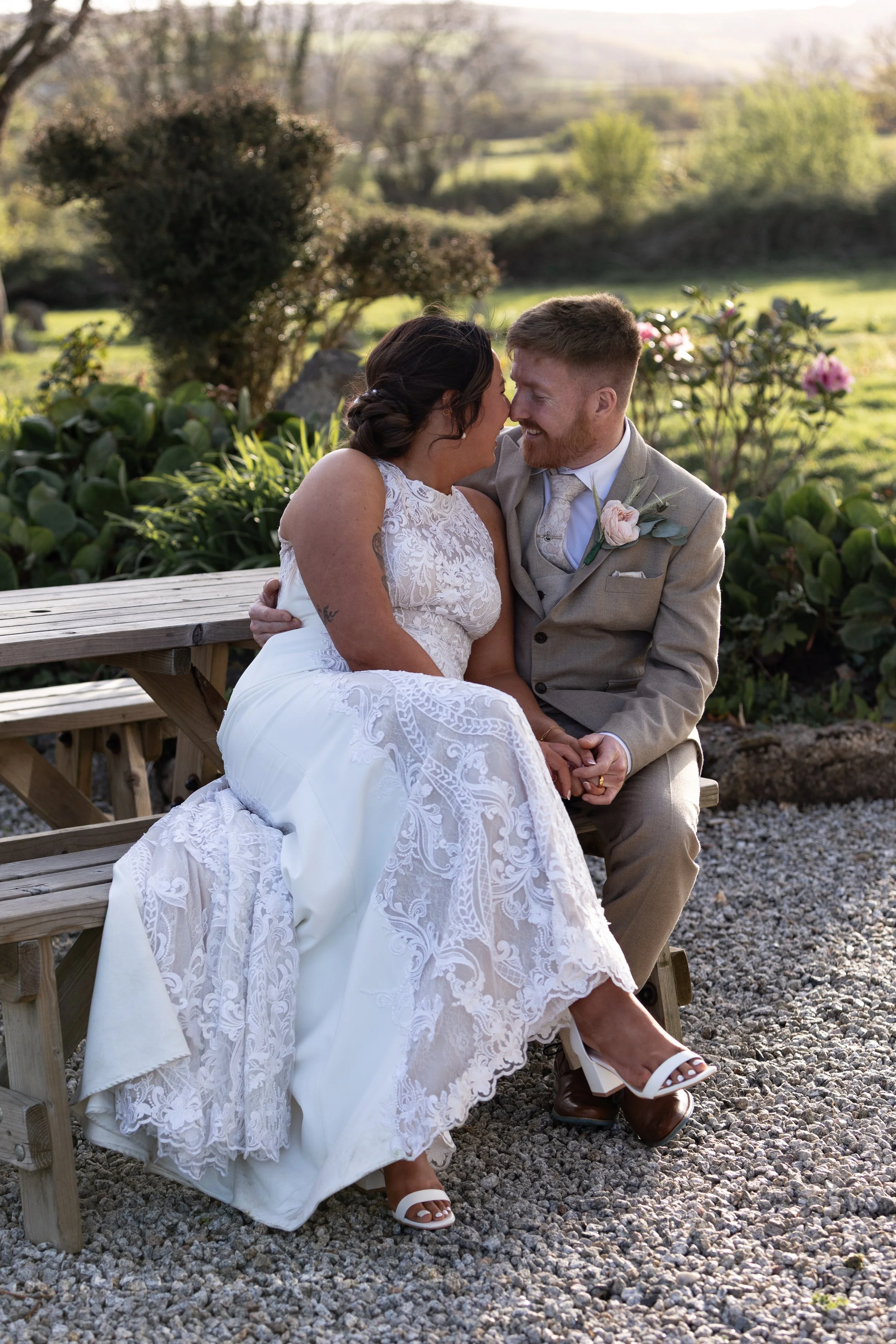 A bride and groom sitting closely and smiling at each other outdoors during a wedding, with lush green landscape in the background.