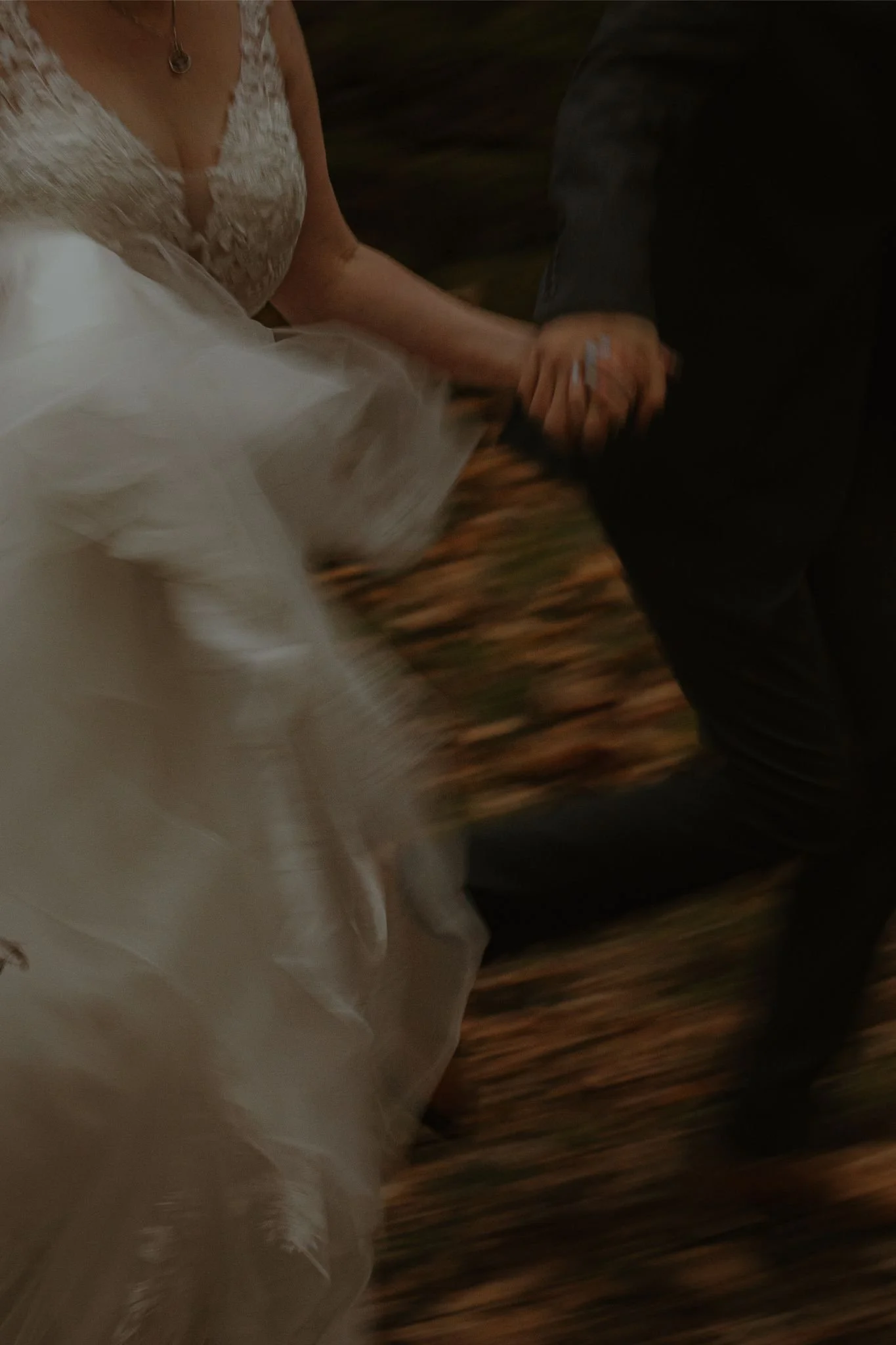 A woman in a wedding dress holding hands with a man dressed in black, dancing together, with a blurred background.