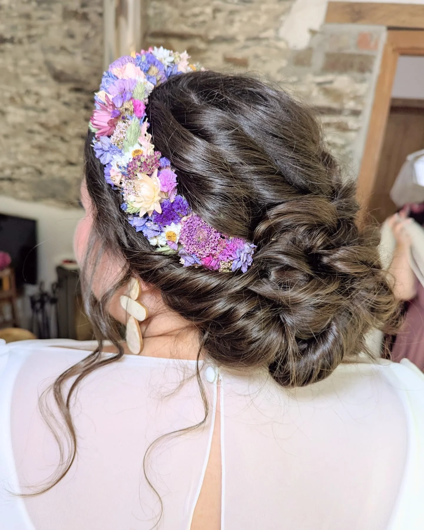 A woman with dark wavy hair styled in an elegant updo, adorned with a colorful floral headband consisting of purple, pink, white, and yellow flowers. She is wearing earrings and a white dress or blouse, with a rustic stone wall background.