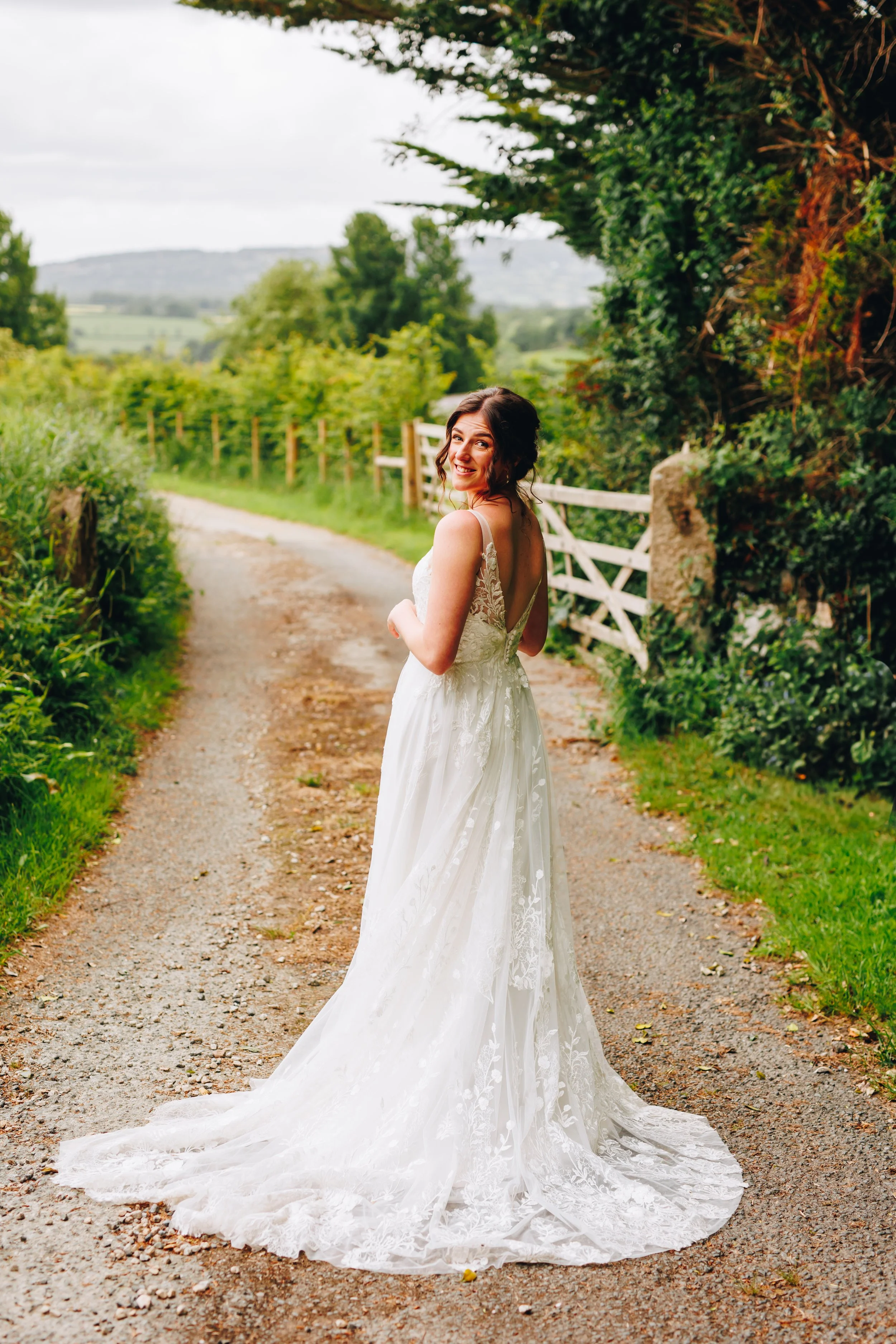 A woman in a white wedding dress standing on a gravel path, smiling and looking back, during daytime in a rural area with green trees and fields in the background.