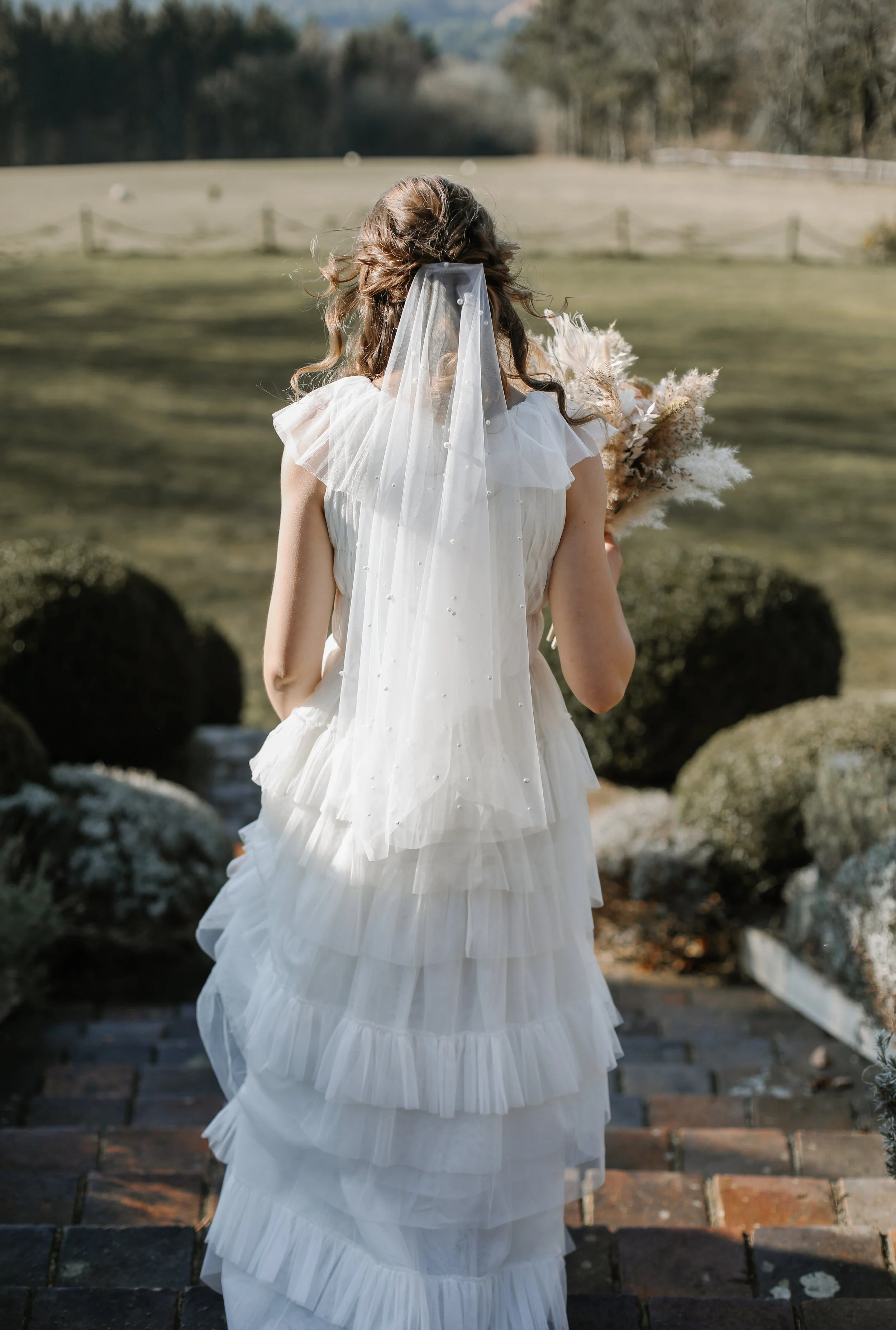 A woman in a white wedding dress with layered ruffles and a veil, holding a bouquet, standing outdoors on brick steps with a grassy field and trees in the background.