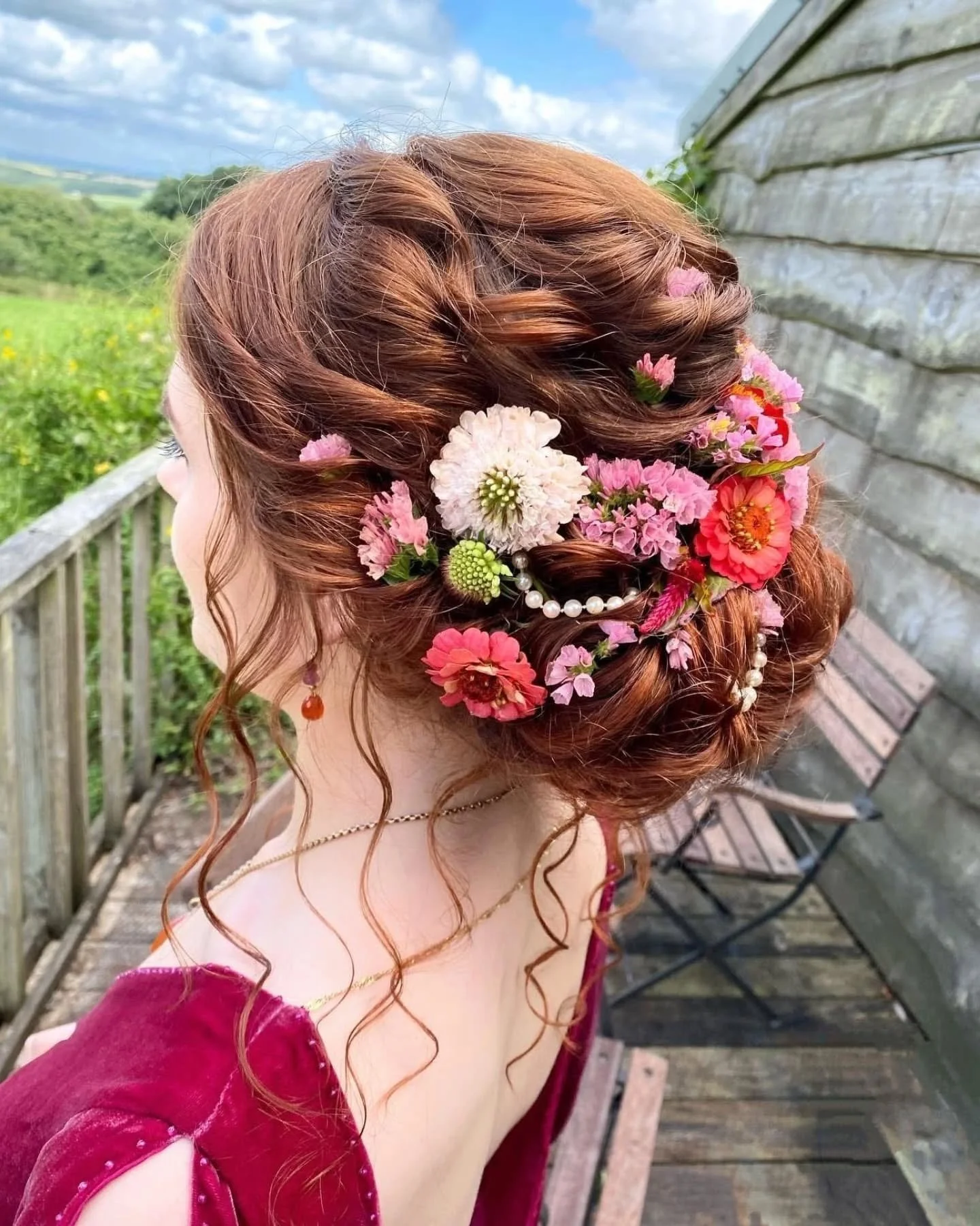 A woman with red hair styled in an updo decorated with pink, white, and red flowers, pearl accents, and curls. She is wearing a burgundy velvet dress and gold jewelry, standing on a wooden balcony overlooking a green landscape with a cloudy sky.