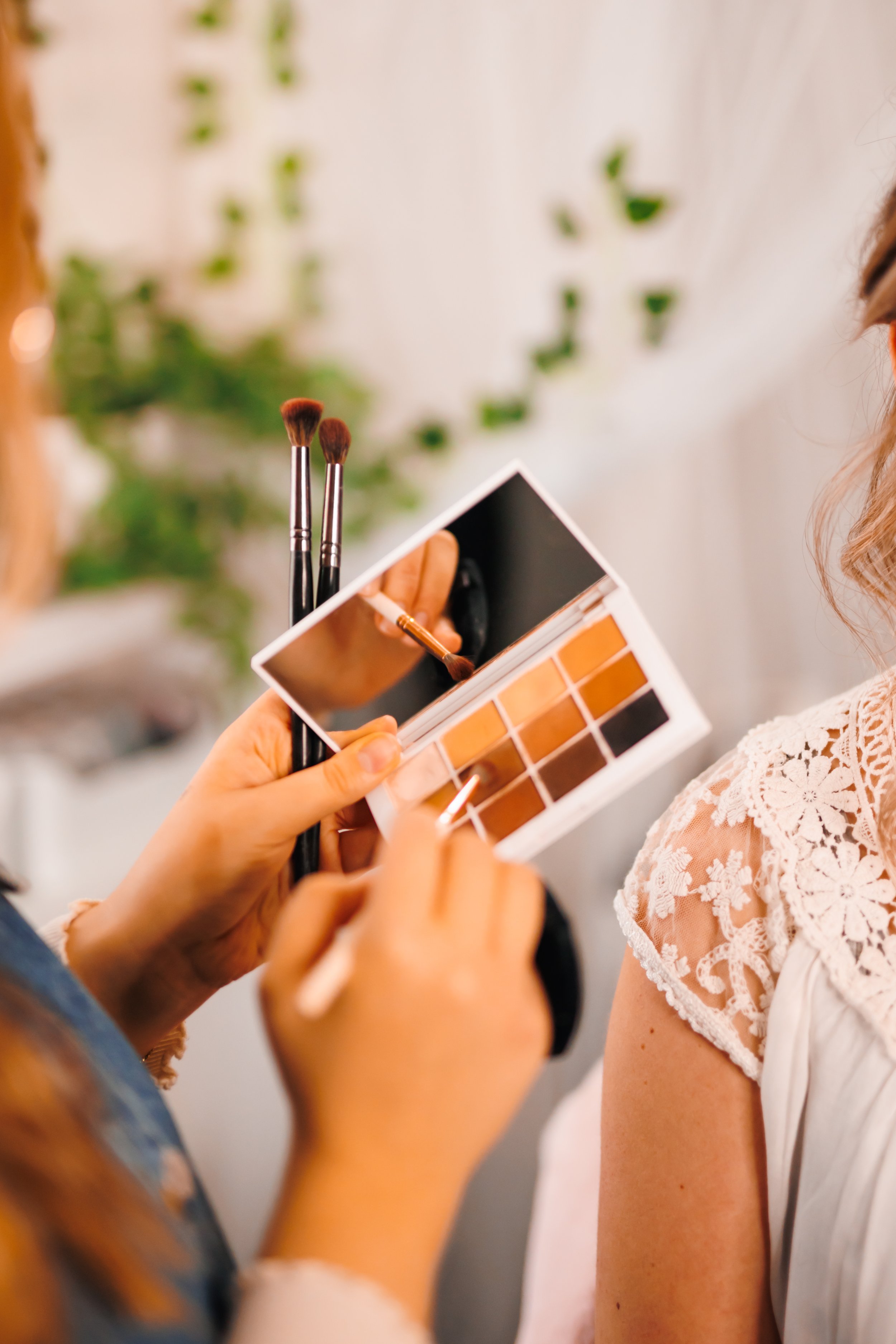Close-up of a person's hands holding a makeup palette and mirror, applying makeup to another person's shoulder, with makeup brushes and a blurred background.