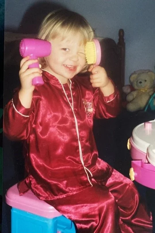 A young girl with blonde hair smiling, holding a pink hairbrush and a pink toy hairdryer, sitting on a pink stool, wearing a red satin princess costume, with a teddy bear and toy makeup set visible in the background.