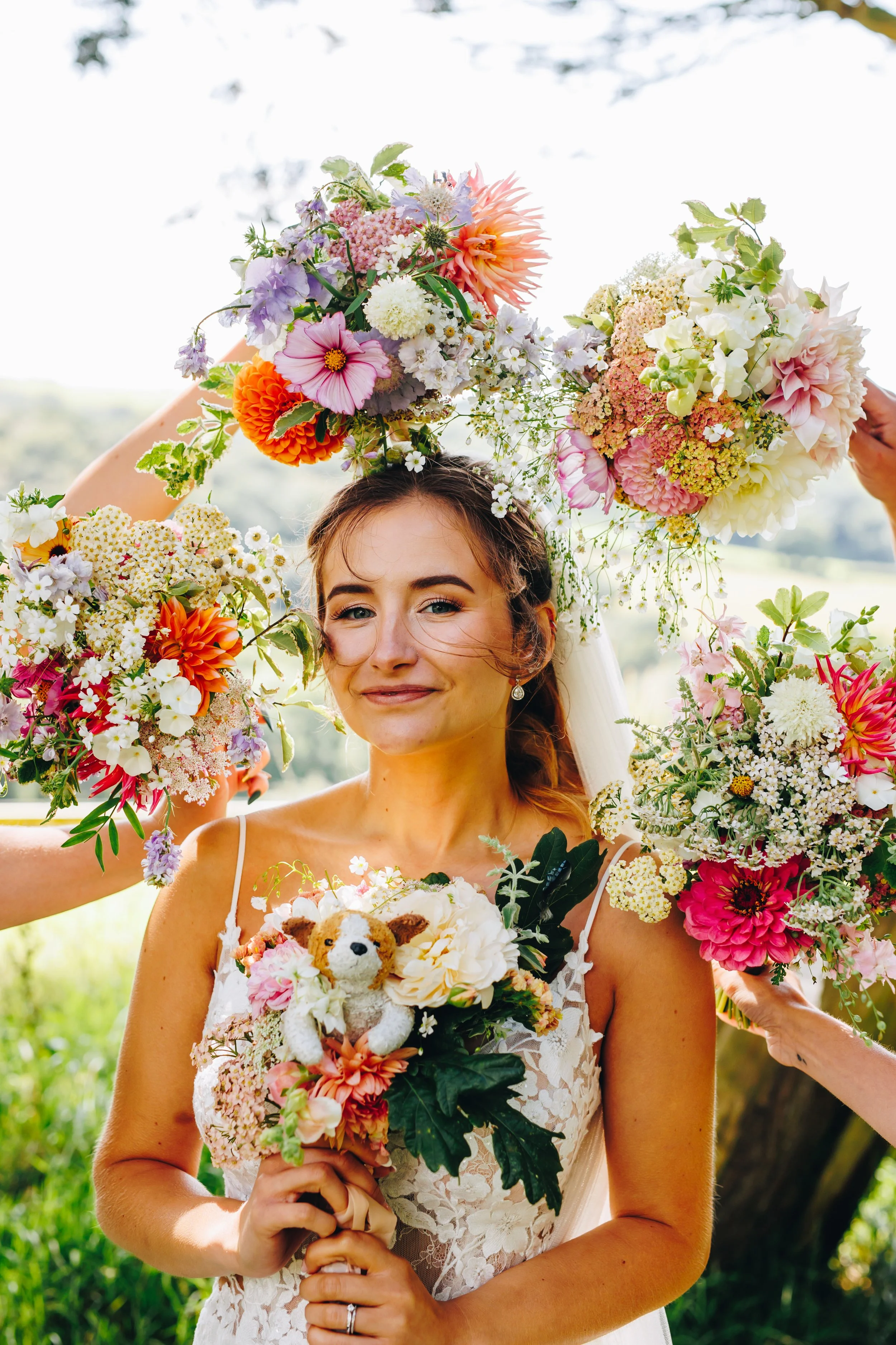 A bride holding a bouquet, surrounded by friends holding large floral arrangements over her head, outdoors during daytime.