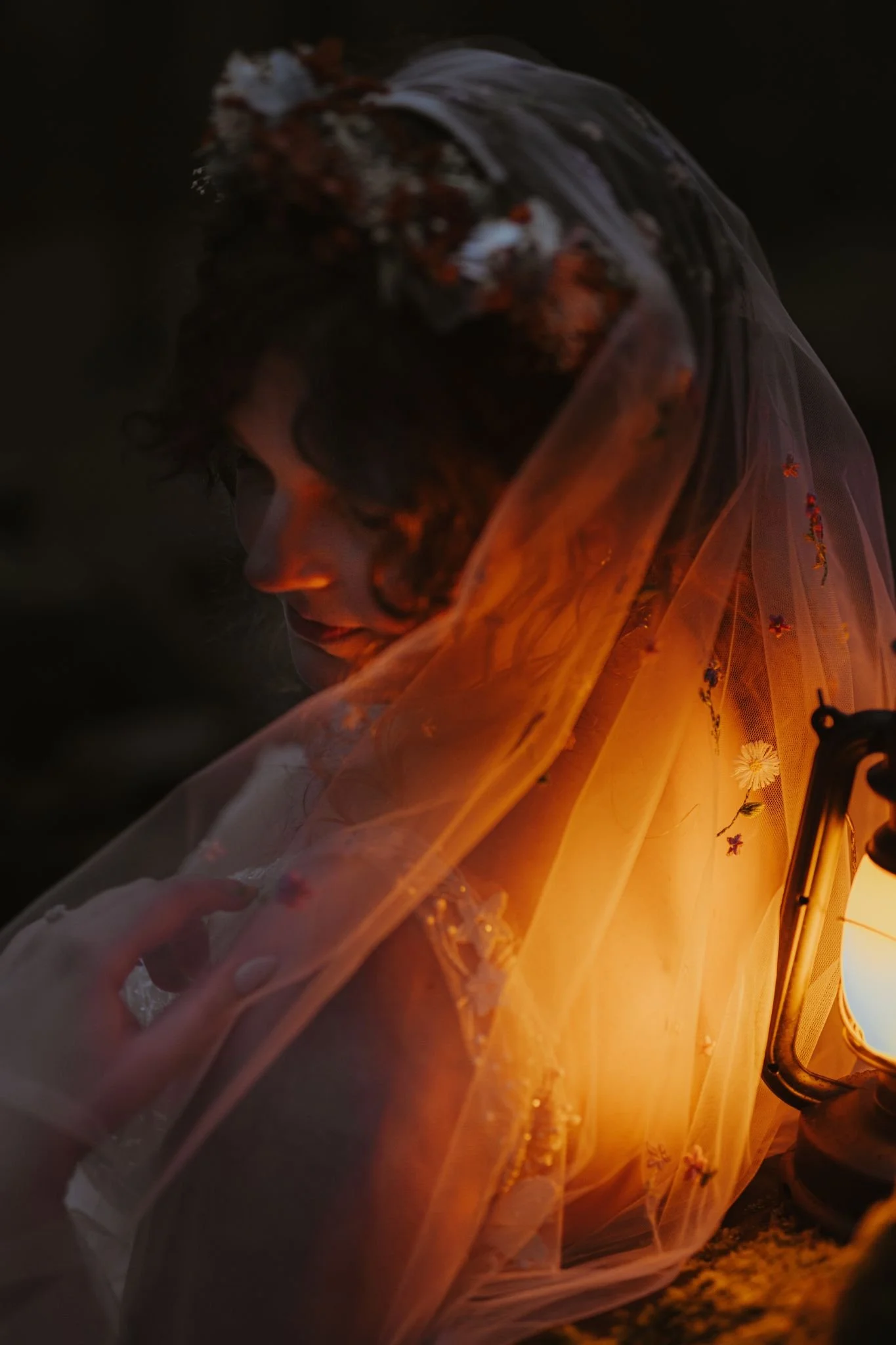 A person with short dark curly hair, wearing a veil with floral embroidery, is sitting close to a lantern, with a warm glow illuminating their face and clothing in a dark setting.