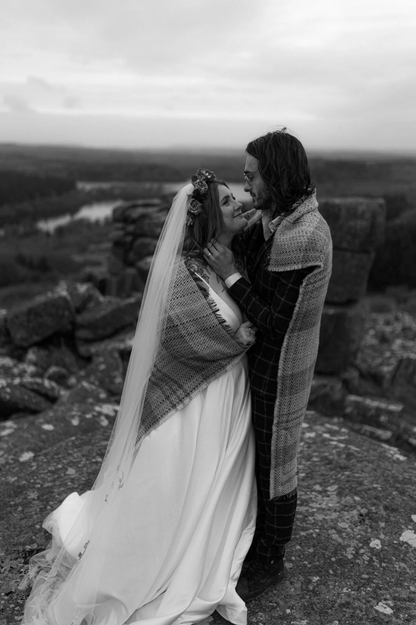 A black and white photo of a couple in wedding attire standing on a rocky outdoor landscape, embraced and gazing into each other's eyes, with a view of a river and hills in the background.