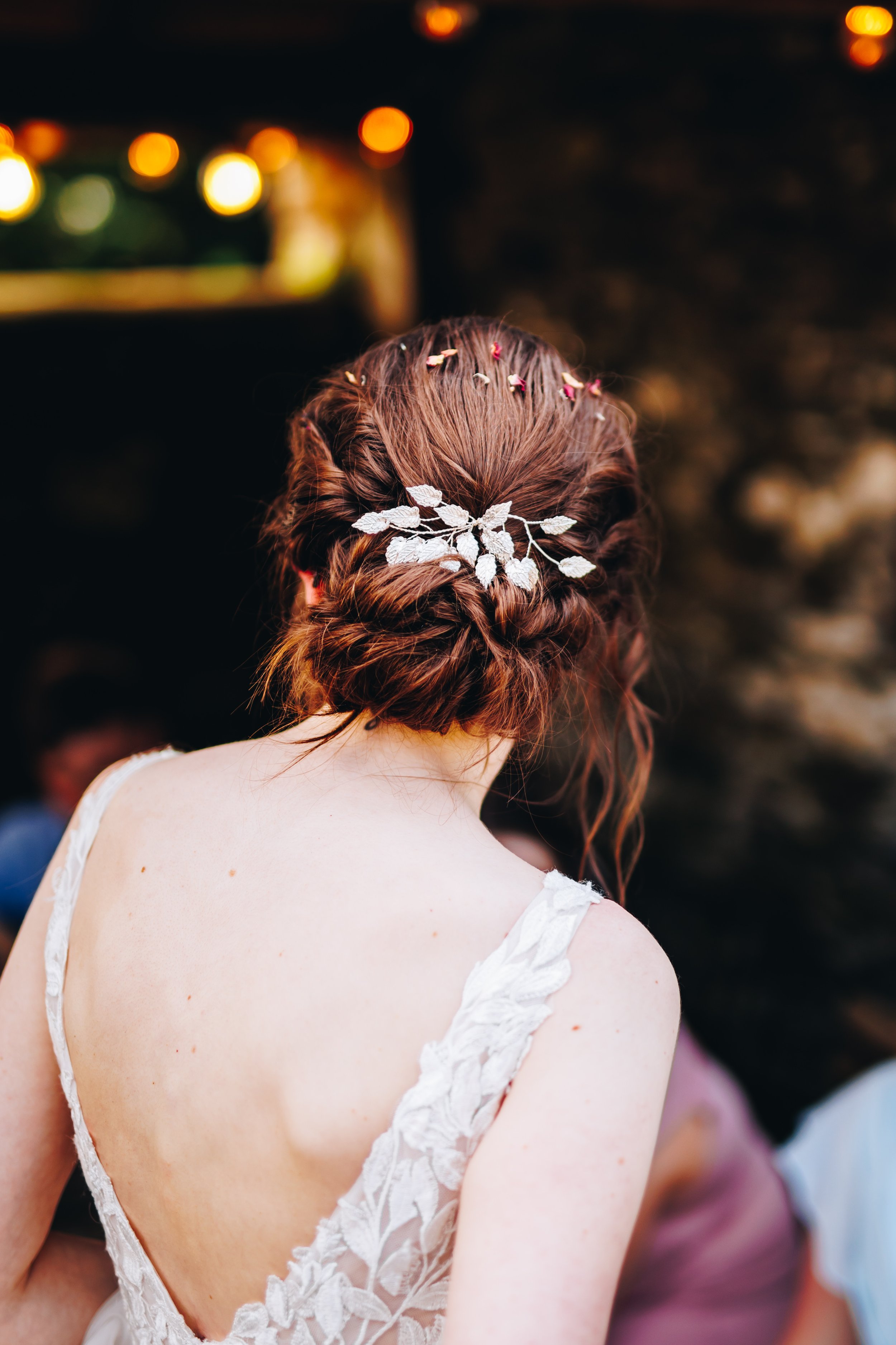 Back view of a woman with red hair styled in an updo, decorated with a floral hairpiece and small flowers, wearing a white lace dress with thin straps.