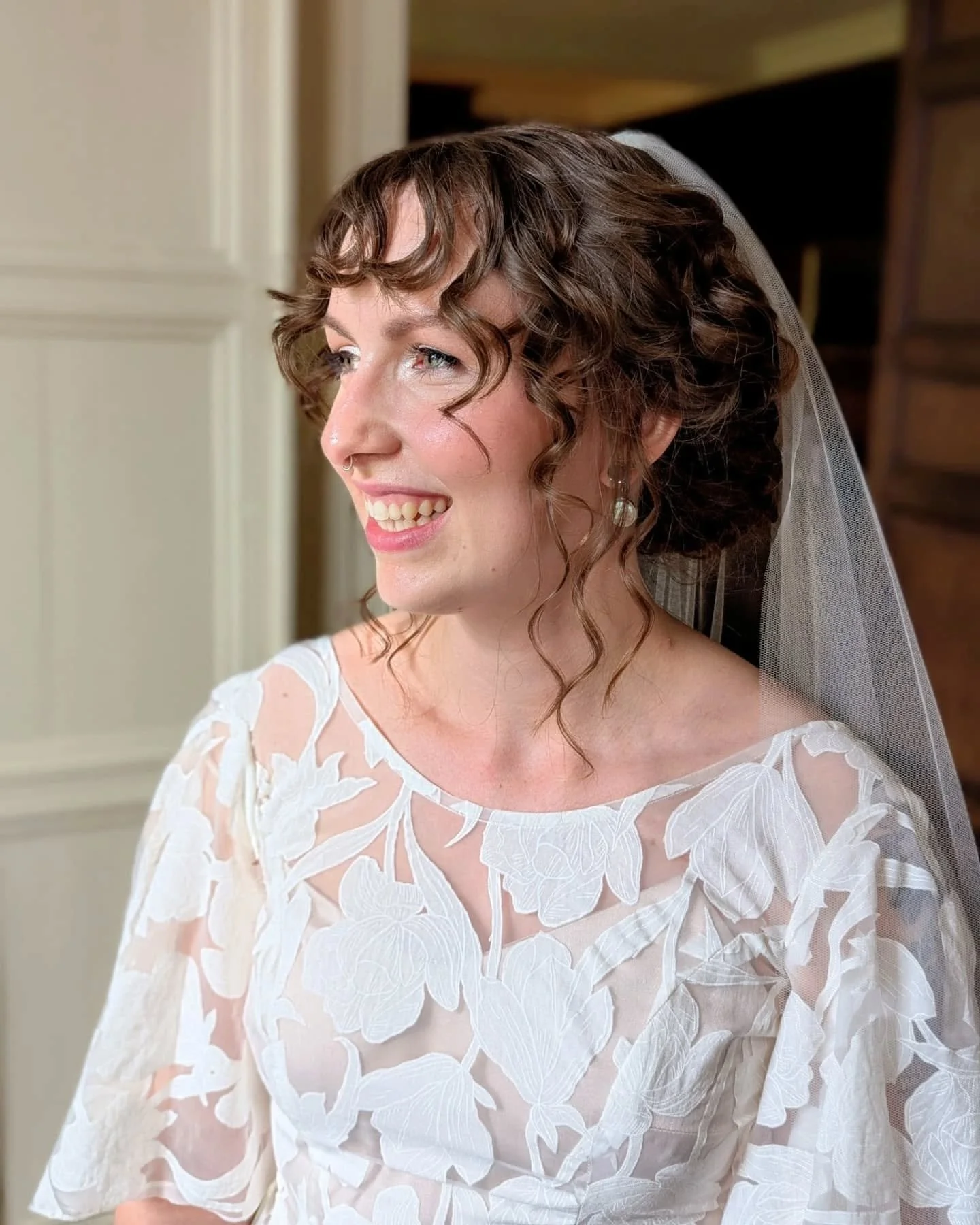A woman with curly brown hair, wearing a wedding veil and a white floral lace dress, smiling and looking to her left.