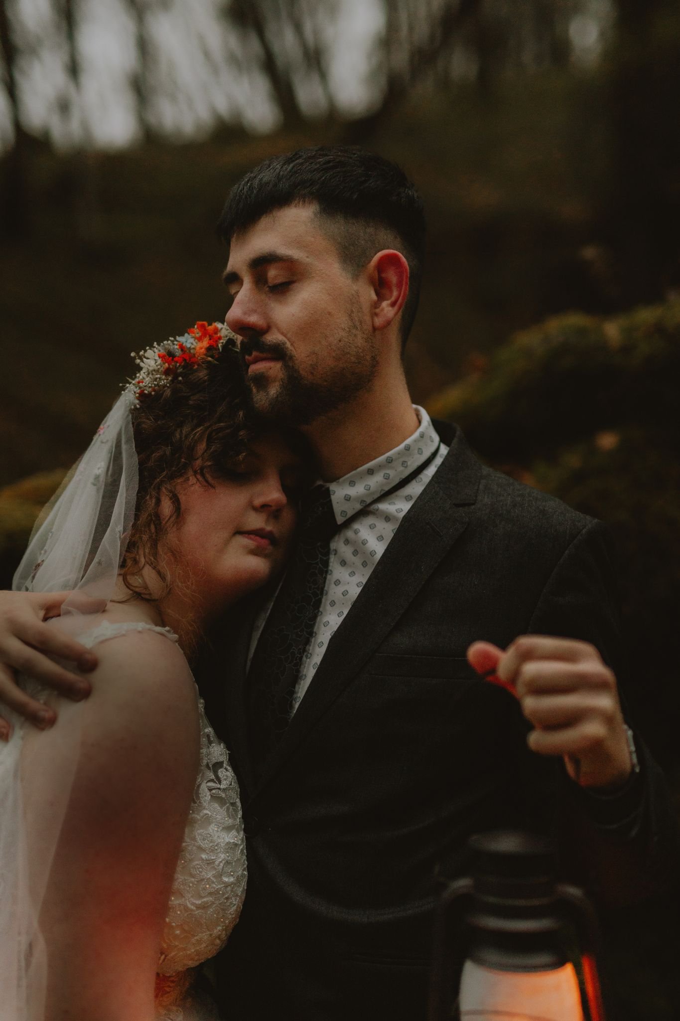 A couple embraces in a forest setting, with the woman wearing a wedding dress and a floral crown, and the man in a suit, both with their eyes closed.