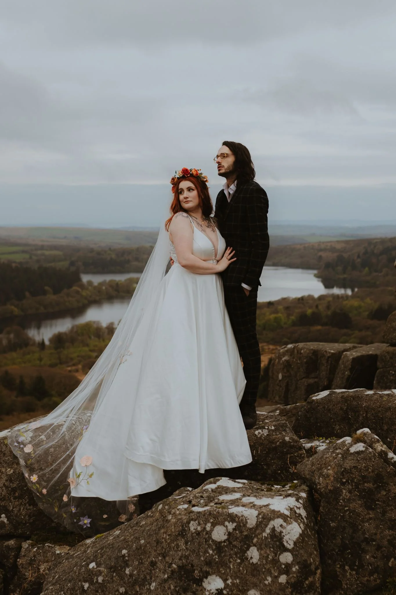A bride and groom standing on rocks outdoors, with a river and landscape in the background during overcast weather.