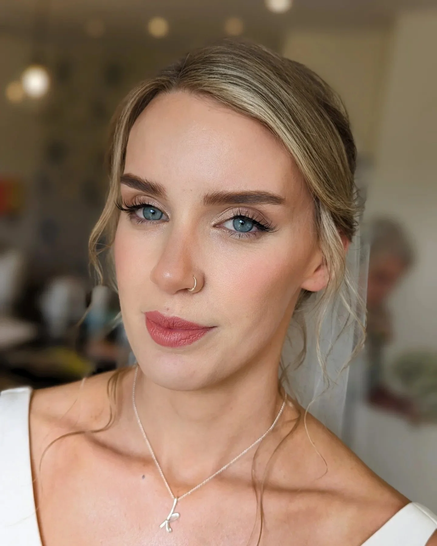 Close-up of a young woman with blonde hair, blue eyes, and a nose ring, smiling softly, wearing a silver necklace with a small pendant.