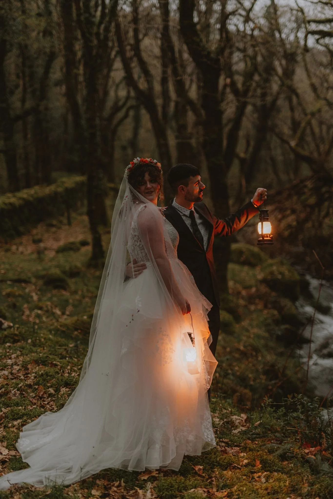 A bride and groom standing outdoors in a wooded area during twilight, holding lanterns. The bride is in a white wedding dress with a veil and floral headpiece, while the groom is wearing a dark suit. The scene is illuminated by the lanterns they are 
