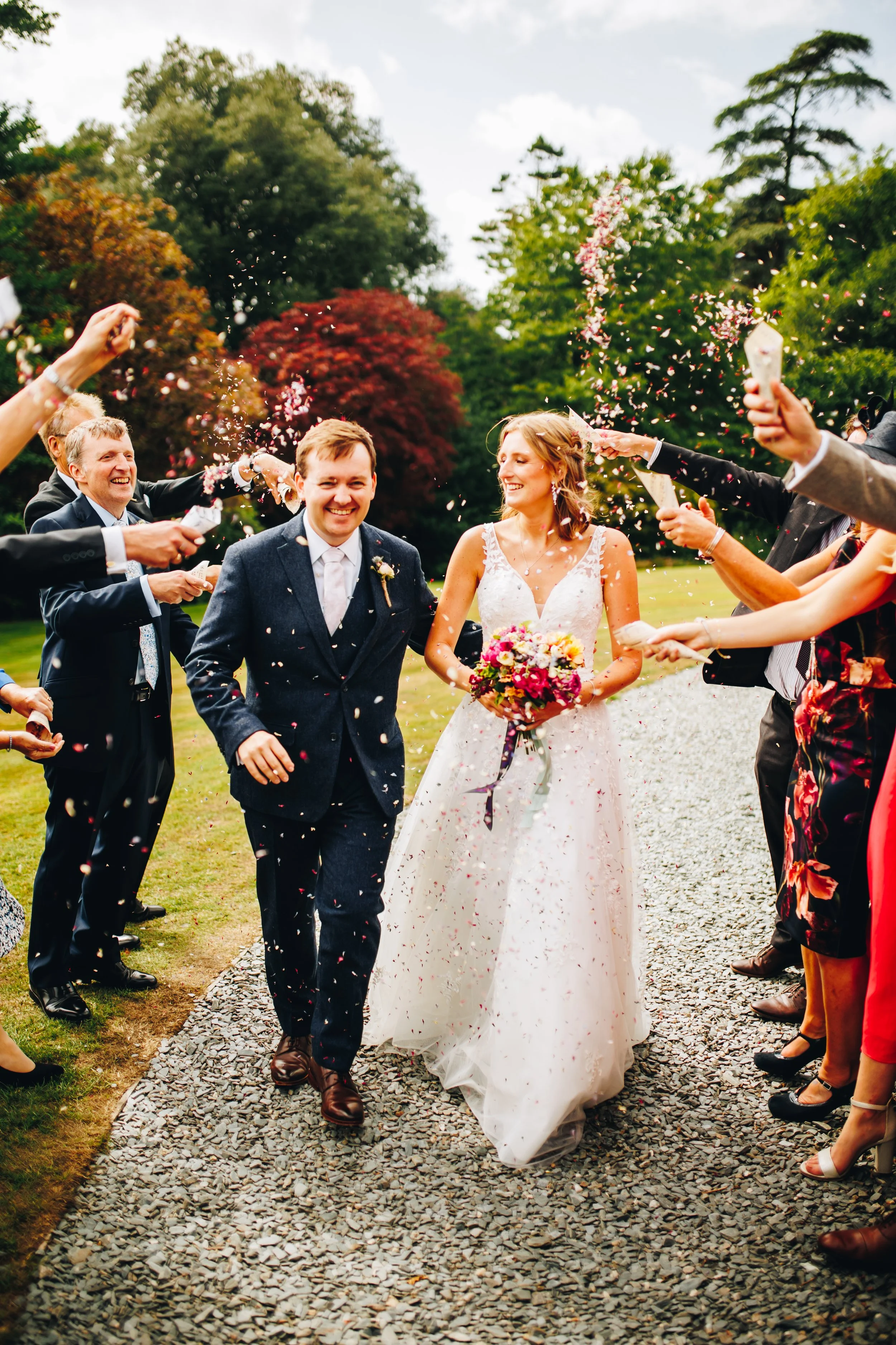 A newlywed couple walking down a path, surrounded by well-dressed friends and family throwing confetti, in a lush green outdoor setting.