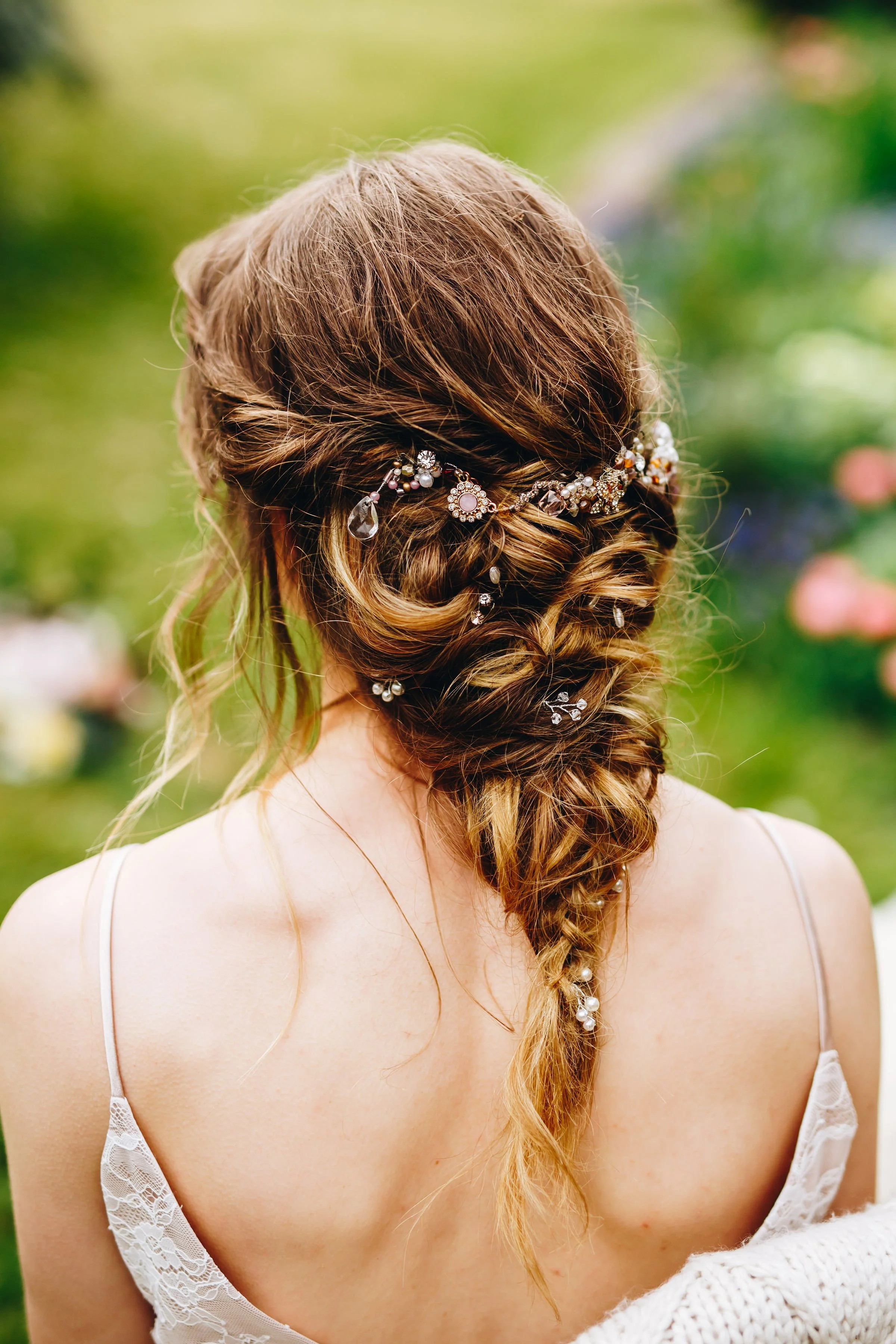 Back view of a woman with styled, braided hair decorated with pearls and rhinestones, wearing a white lace dress, outdoors with blurred greenery in the background.
