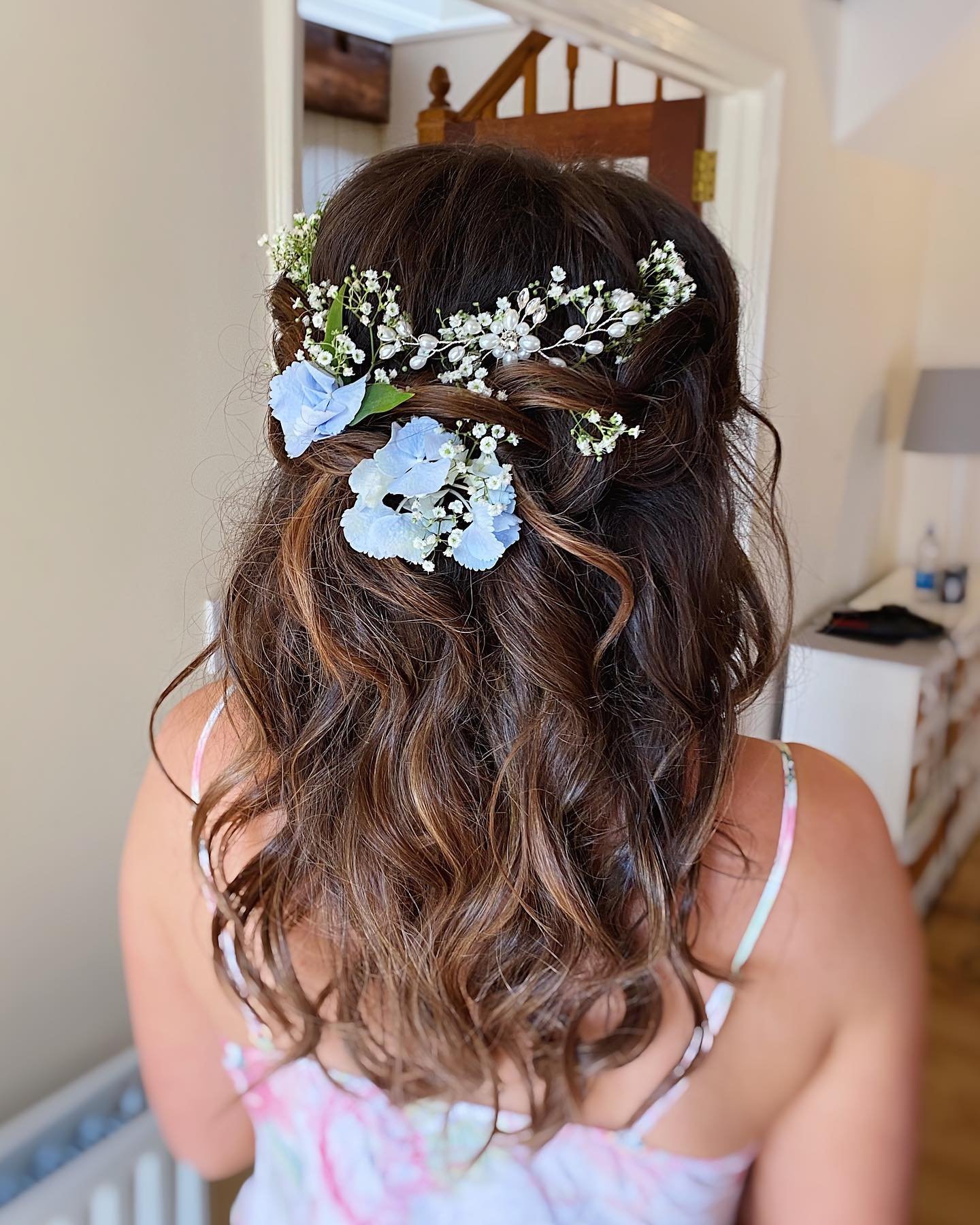 Back view of a woman with wavy brown hair decorated with a floral hairpiece made of white flowers, baby’s breath, and light blue flowers, wearing a pastel-colored dress with thin straps.