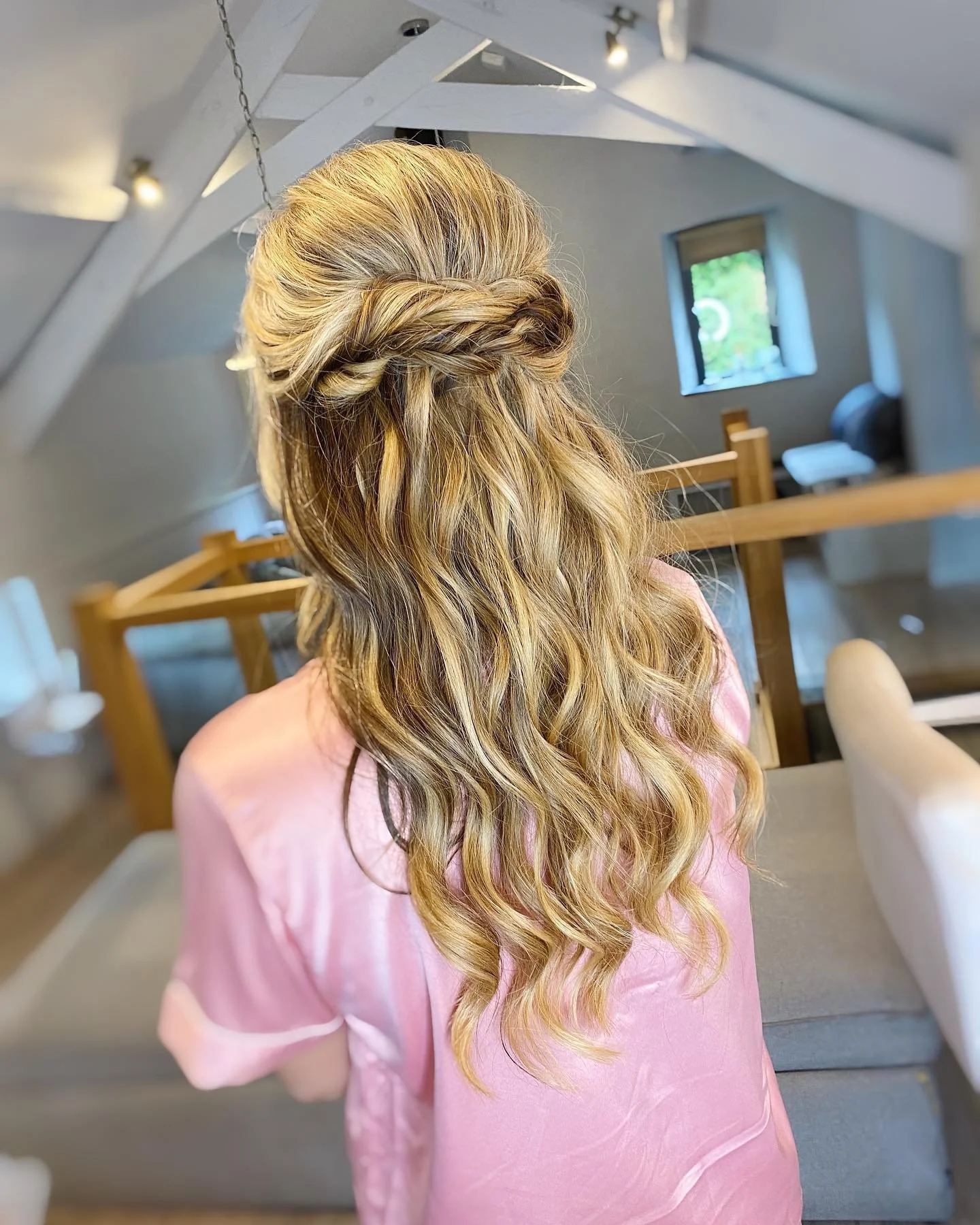 Back view of a woman with long, wavy blonde hair styled with a twisted braid, wearing a pink satin robe, inside a cozy room with sloped ceiling and windows.