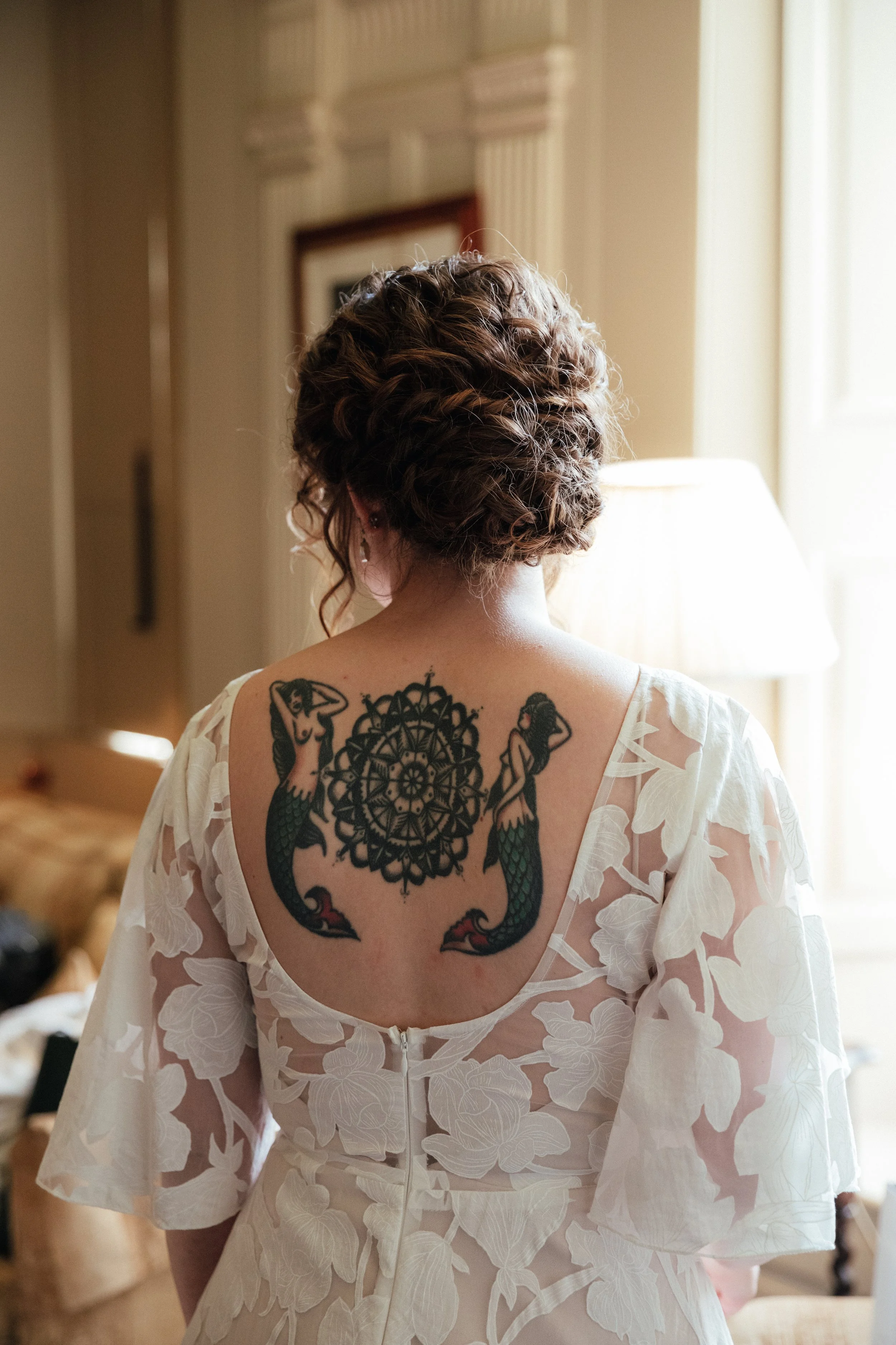 Back of a woman with curly hair wearing a white dress with floral patterns, showing a tattoo of two mermaids and a decorative circular design on her back, indoors with a lamp and picture frame in the background.