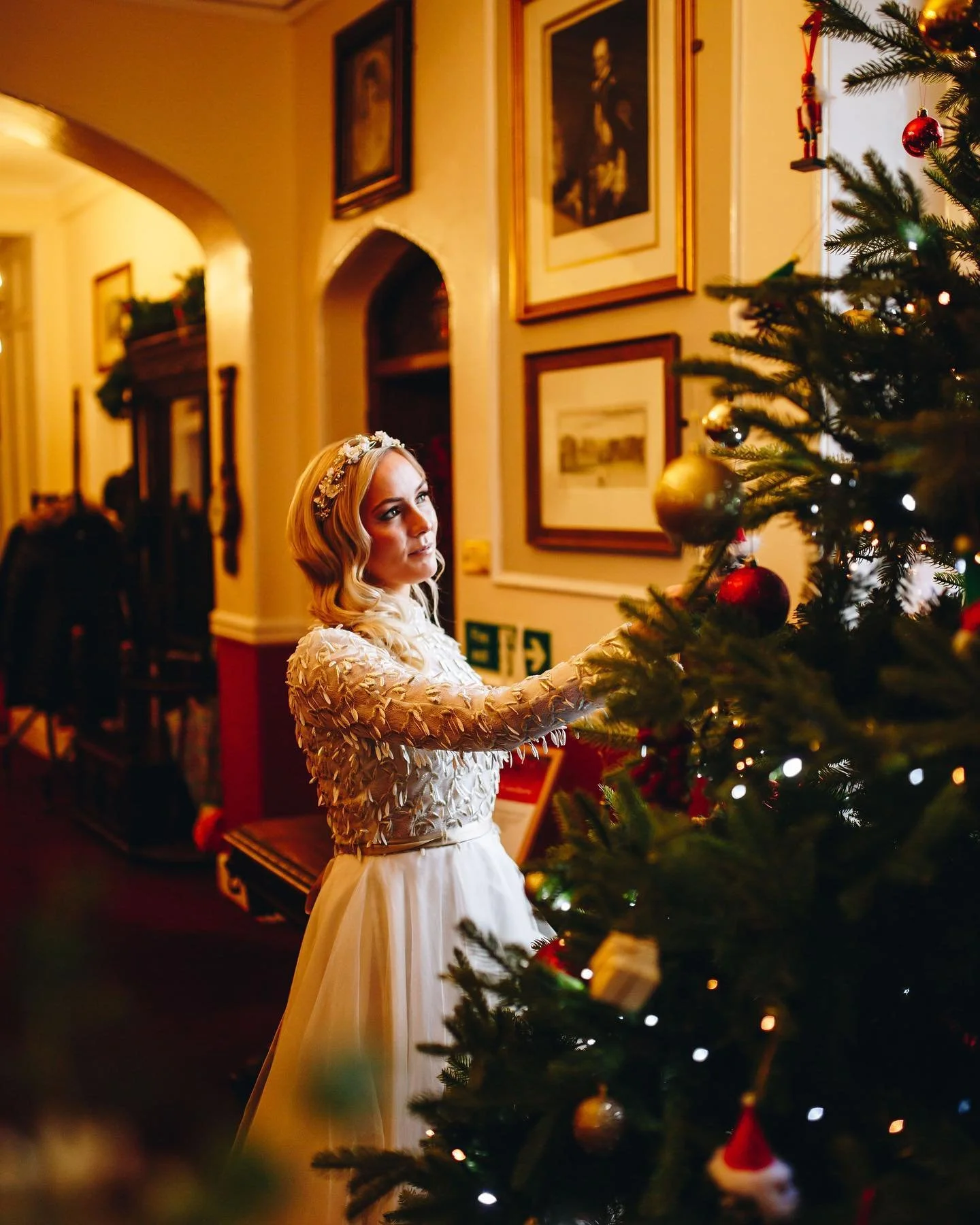 A young woman in a white dress and floral headband decorates a Christmas tree in a cozy, warmly lit room with framed pictures on the wall.