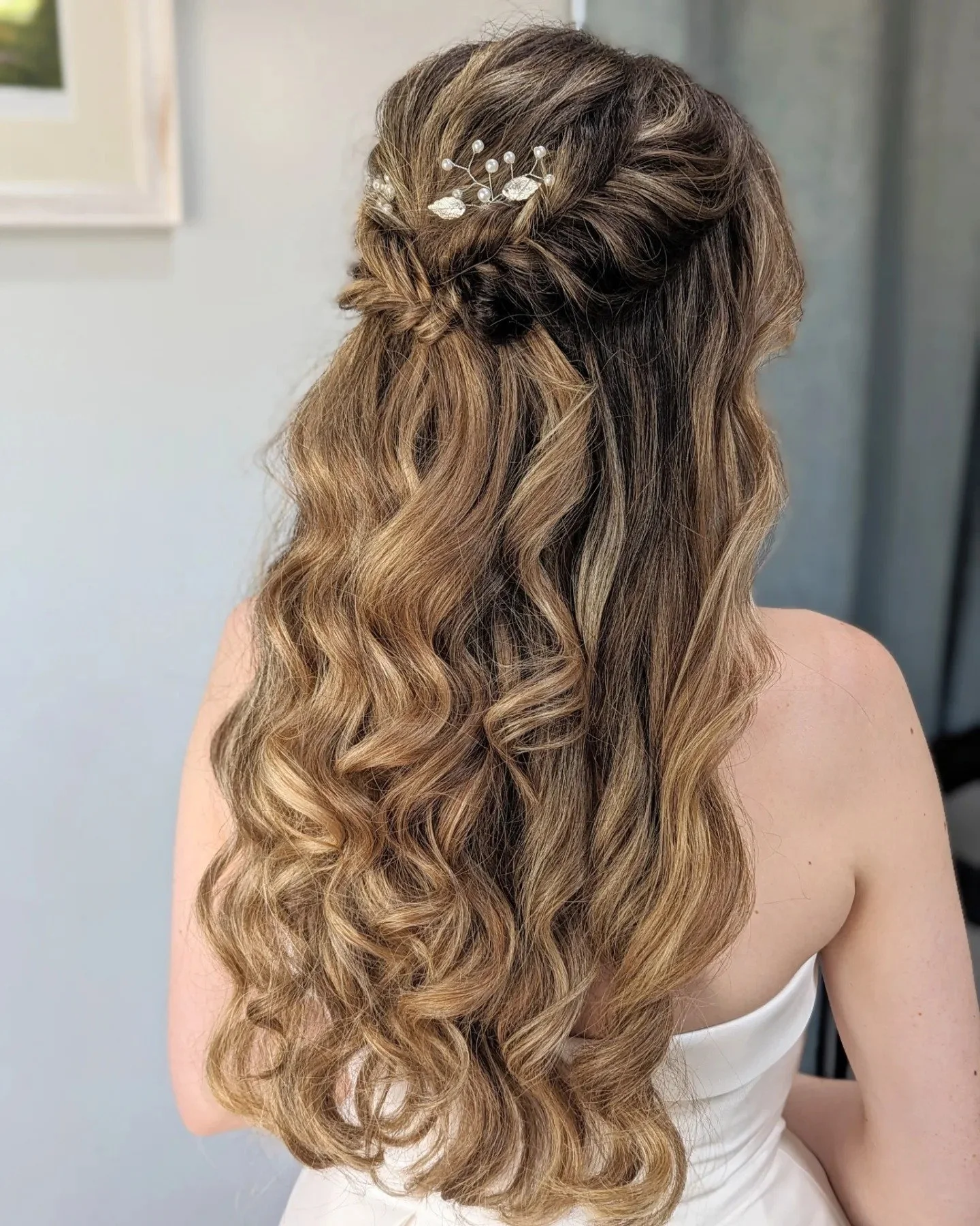 Back view of a woman with long, wavy, blonde hair styled with a twisted half-updo and a decorative hair accessory with pearls and leaves.