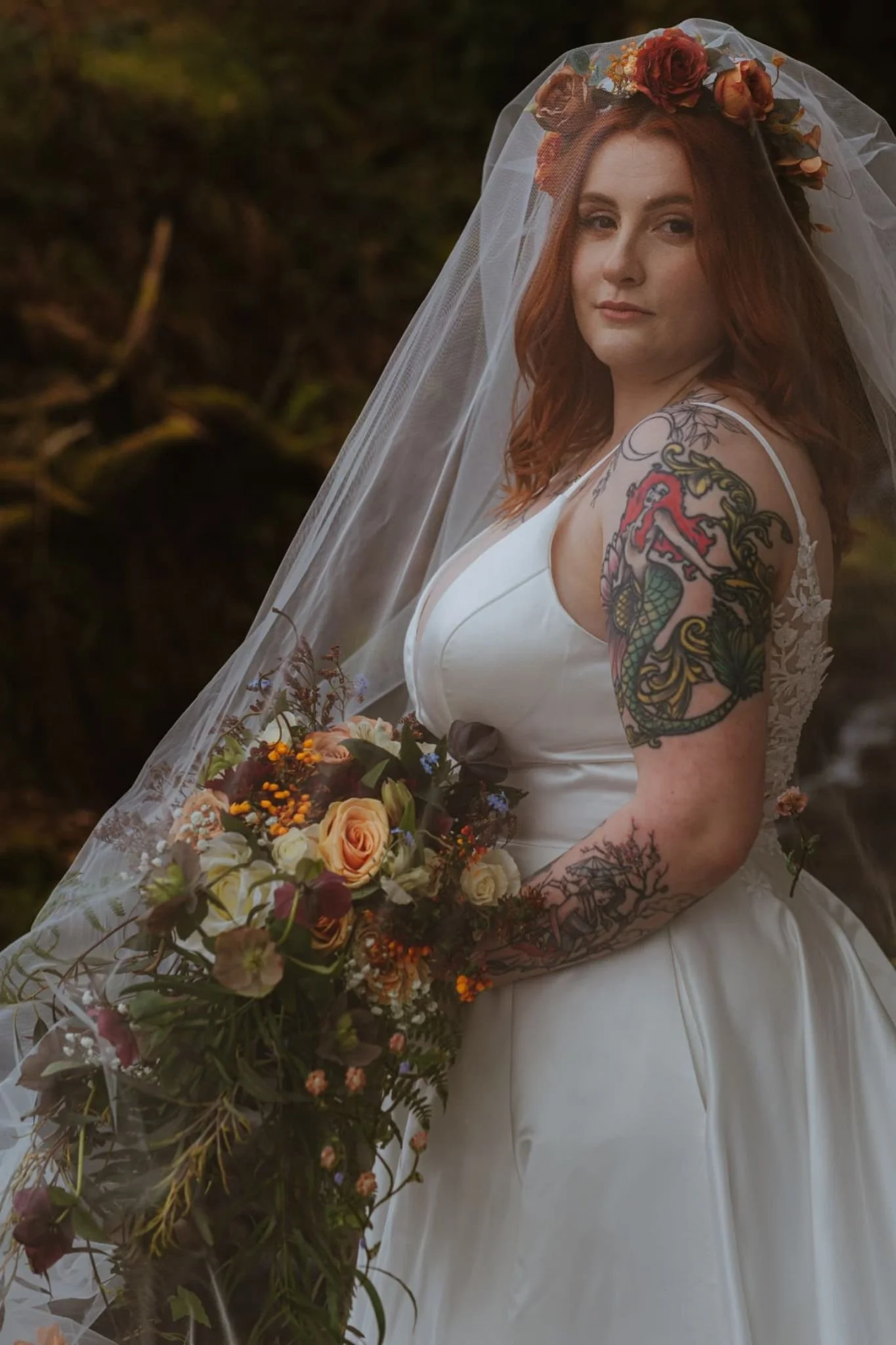 Woman with long red hair in a white wedding dress, wearing a flower crown and veil, holding a bouquet of flowers, outdoors with a dark background.