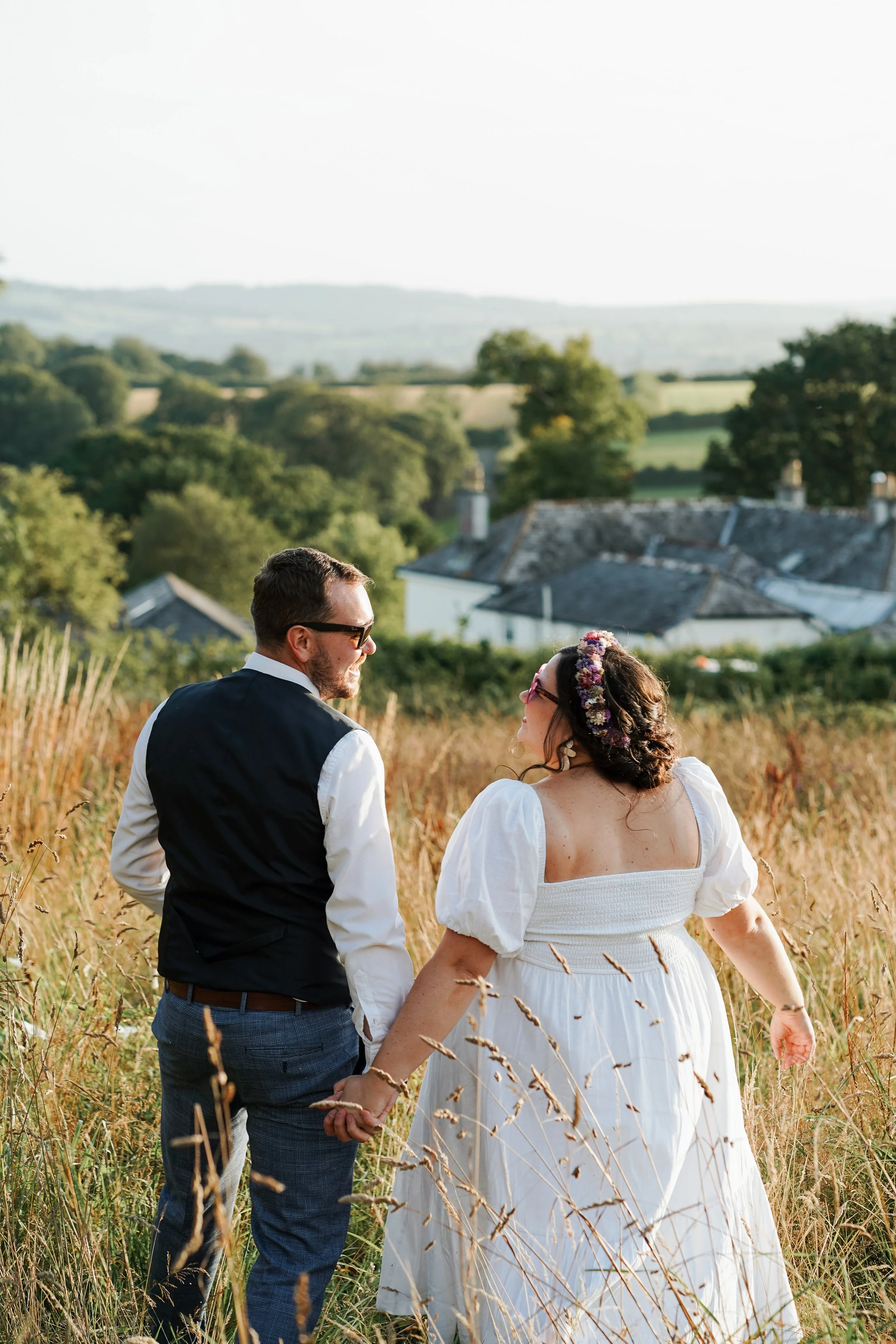 A couple holding hands, walking through a grassy field with a rural village in the background, during sunset.